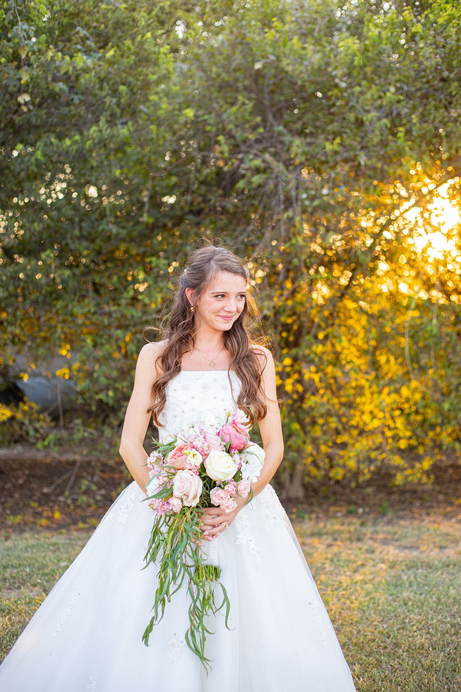 Bride in white ballgown holds pink floral bouquet against autumn foliage backdrop.