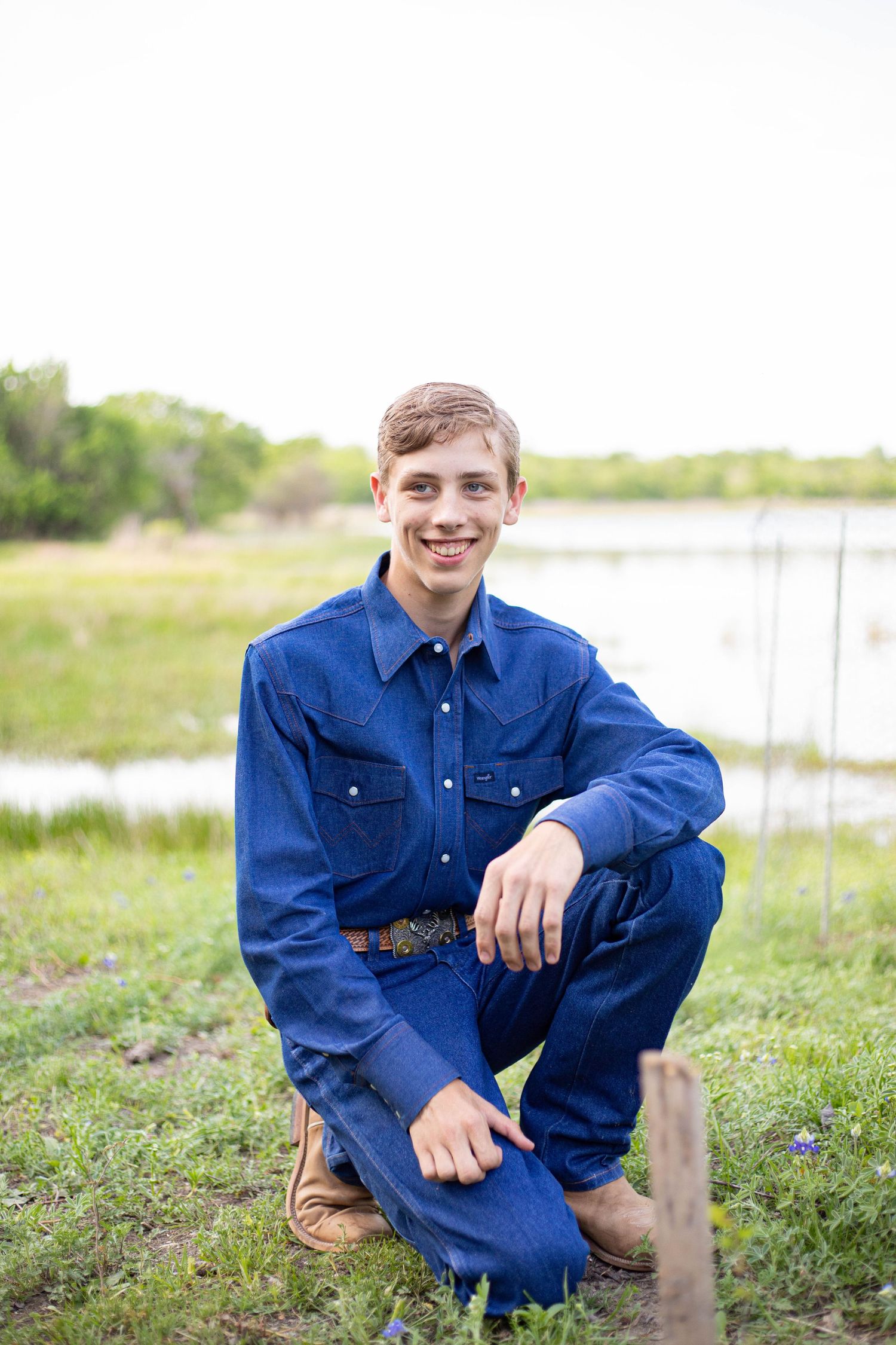 A smiling teenage boy in a blue denim outfit kneels on green grass near a pond in a rural outdoor setting.