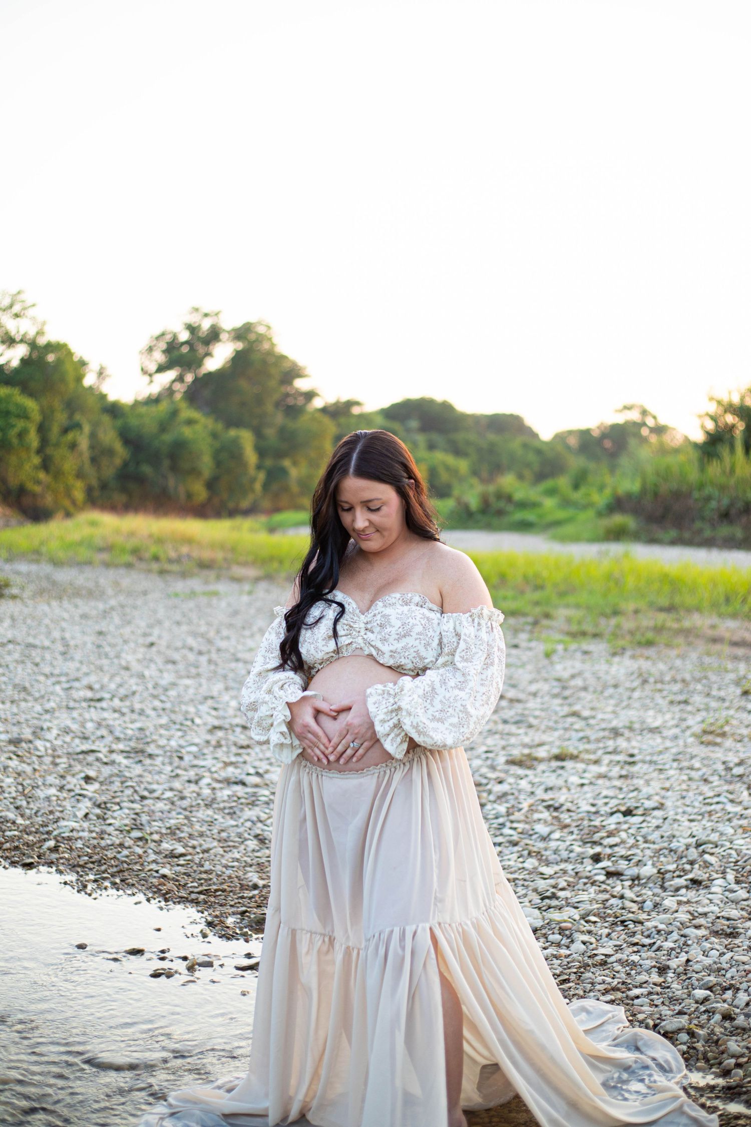 Pregnant woman in white lace off-shoulder gown cradling baby bump outdoors near a rocky shoreline at golden hour.