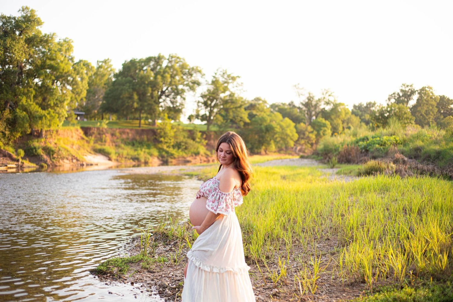 Pregnant woman in white skirt and floral top poses by a sunlit river surrounded by lush green grass and trees.