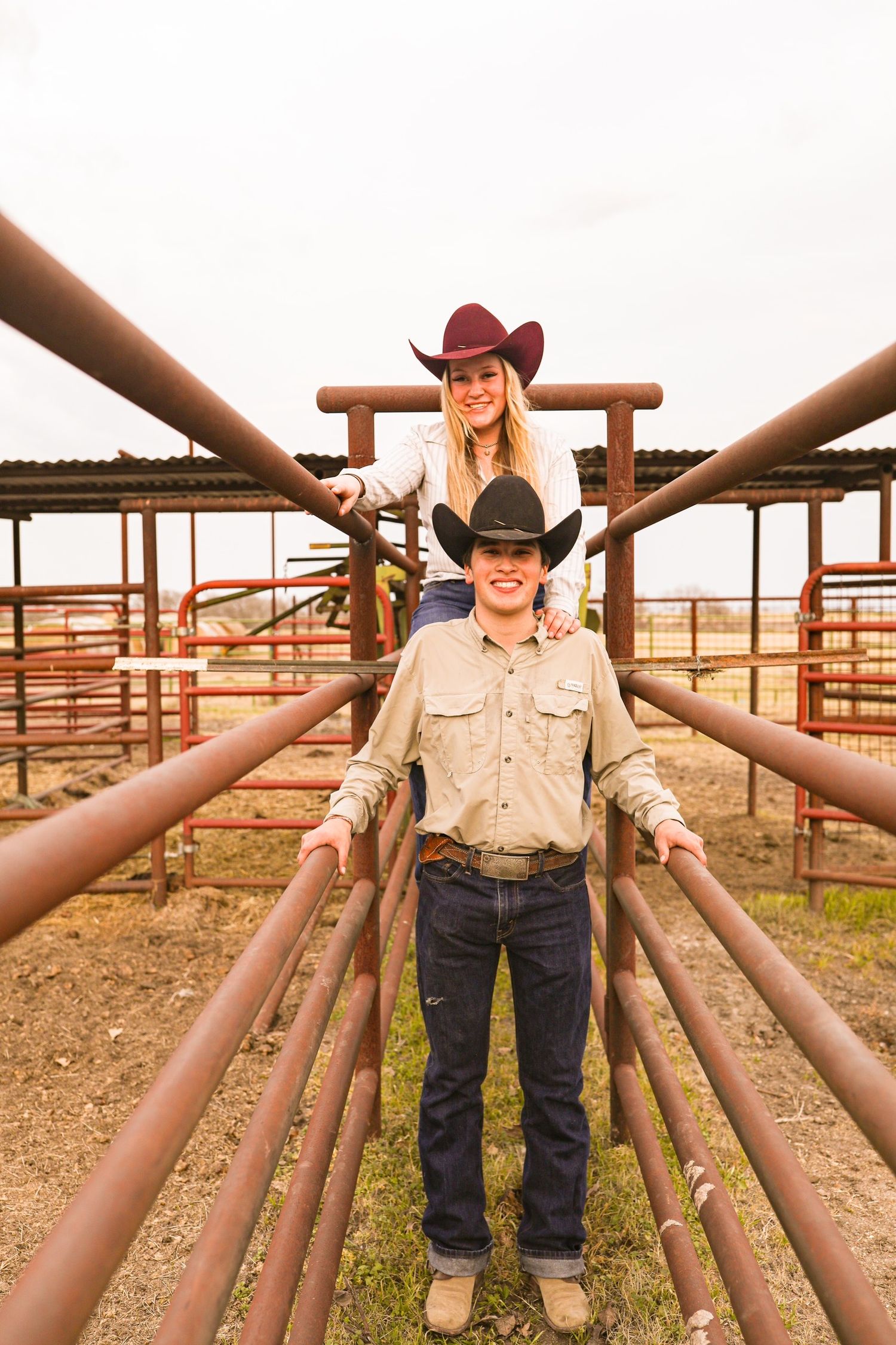 Couple poses on rustic metal cattle chute at ranch wearing western attire.