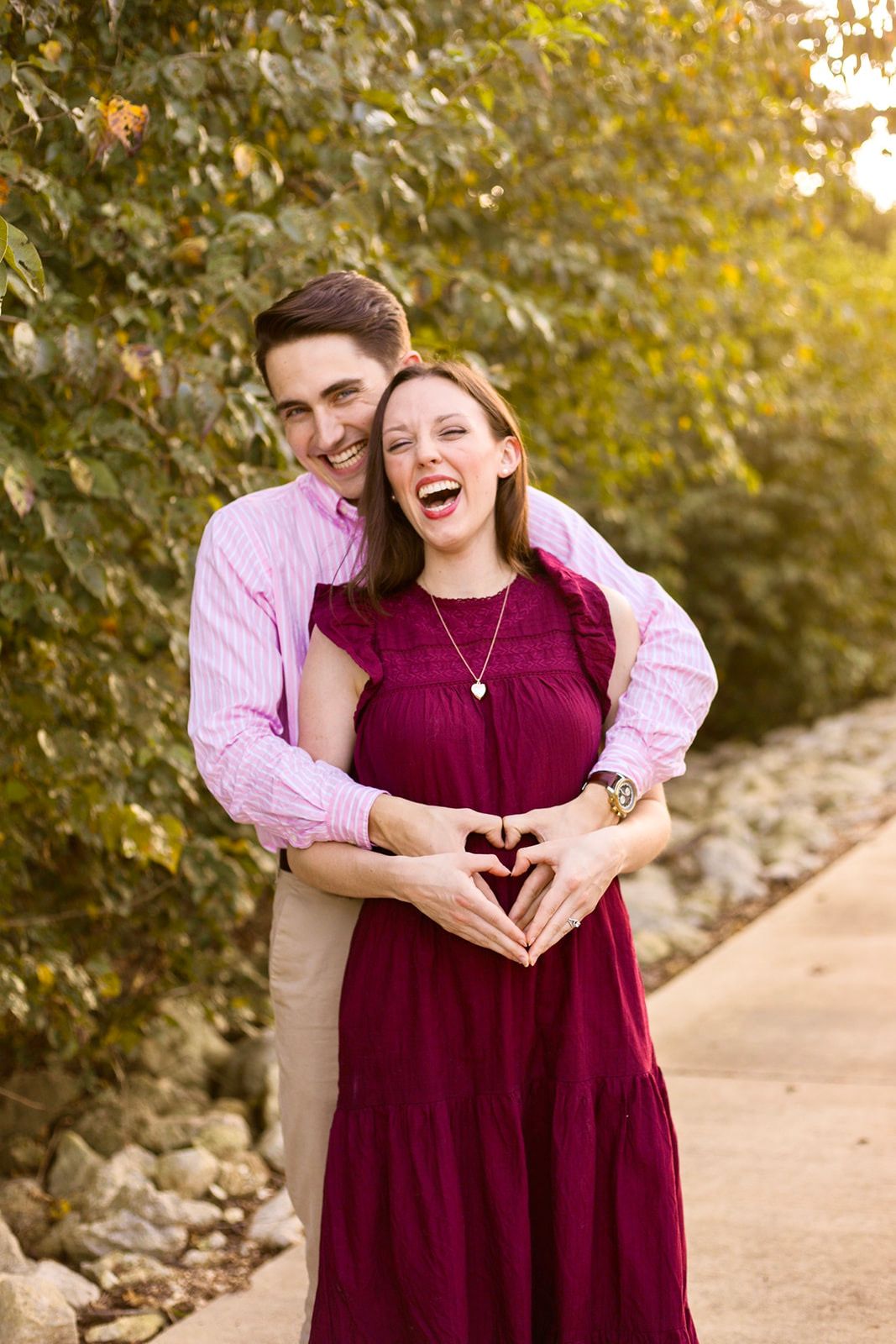 Happy couple making a heart shape with hands over pregnant belly outdoors, surrounded by lush green foliage.