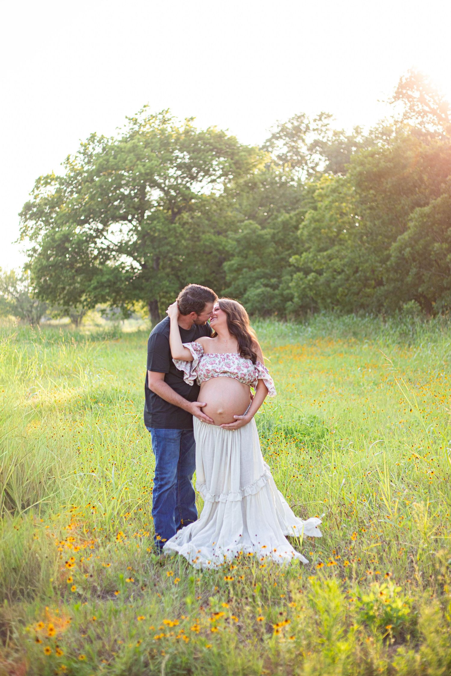 Couple sharing a tender moment in a sunlit meadow, pregnant woman in white gown cradling her baby bump.