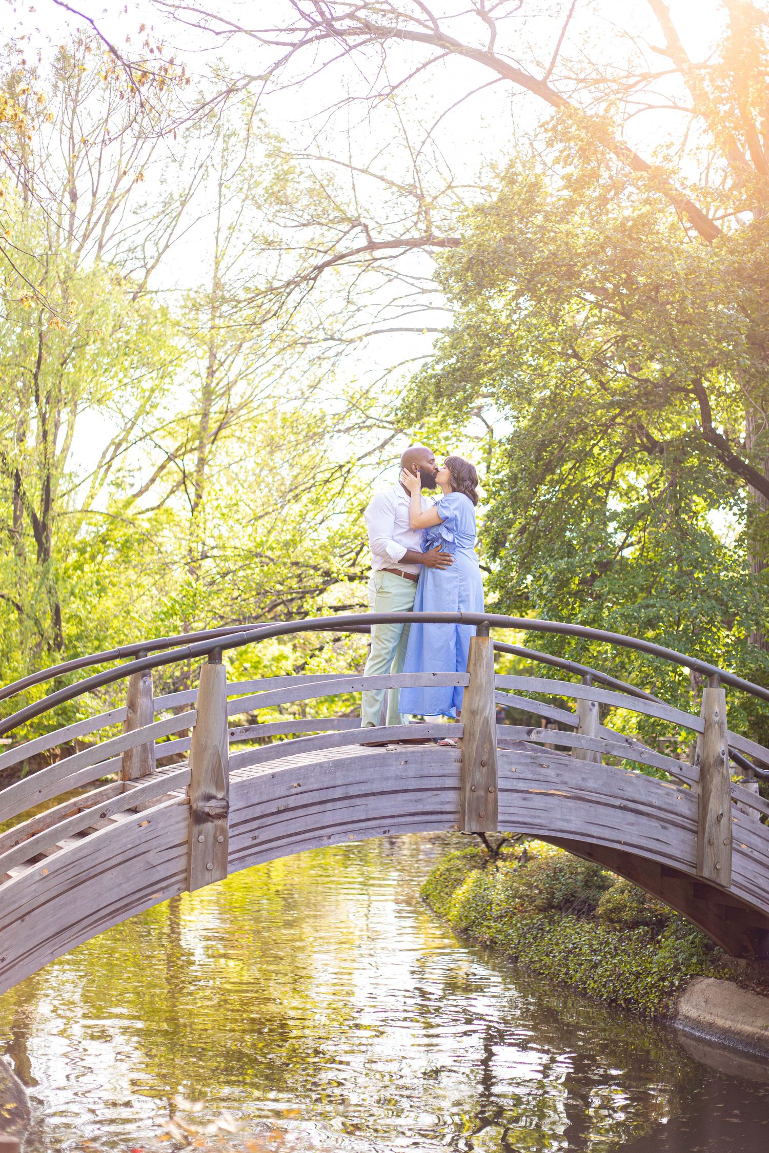 Couple standing on arched wooden bridge over pond during golden hour in garden.