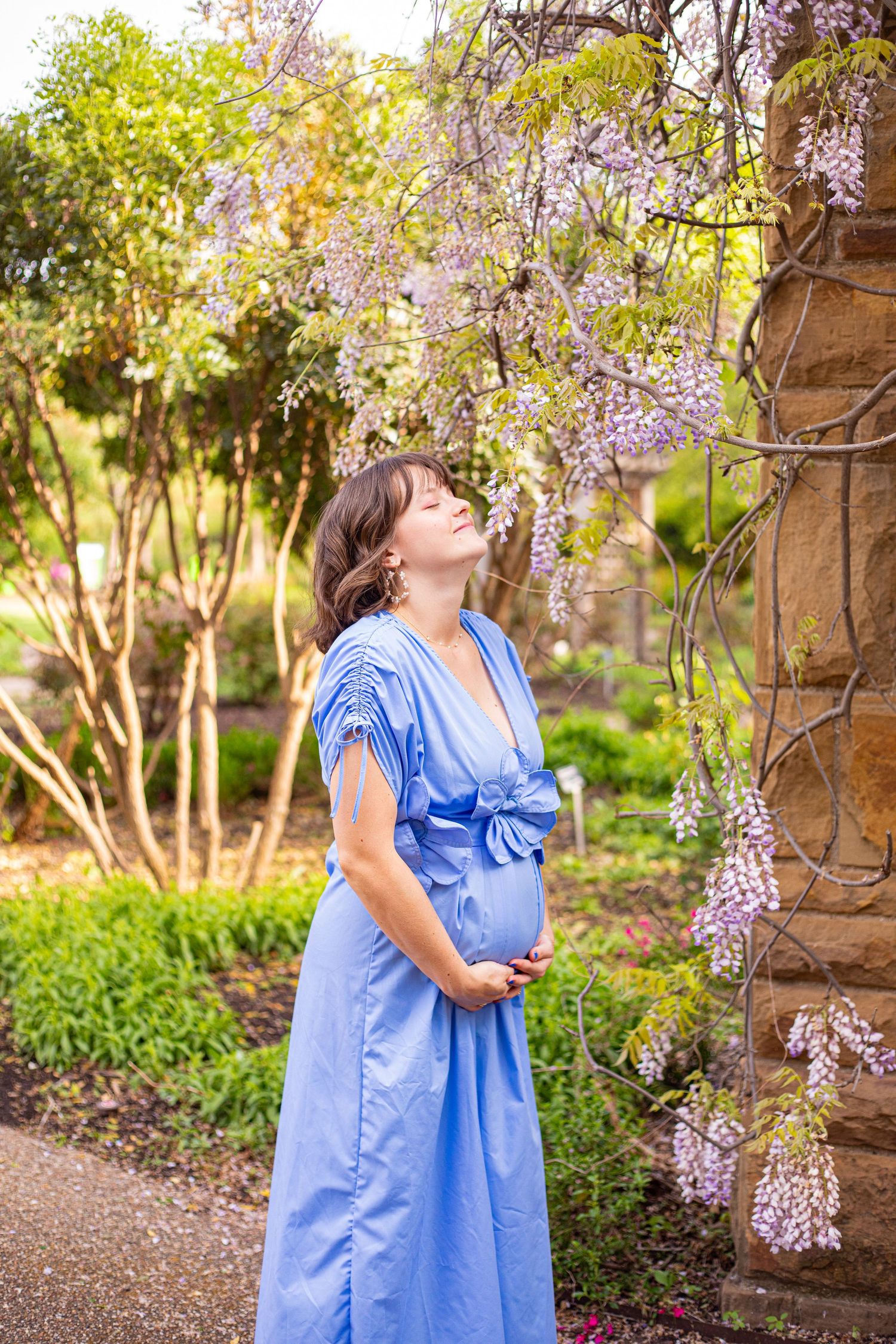 Expectant mother in flowing blue dress poses under blooming wisteria in a garden setting.