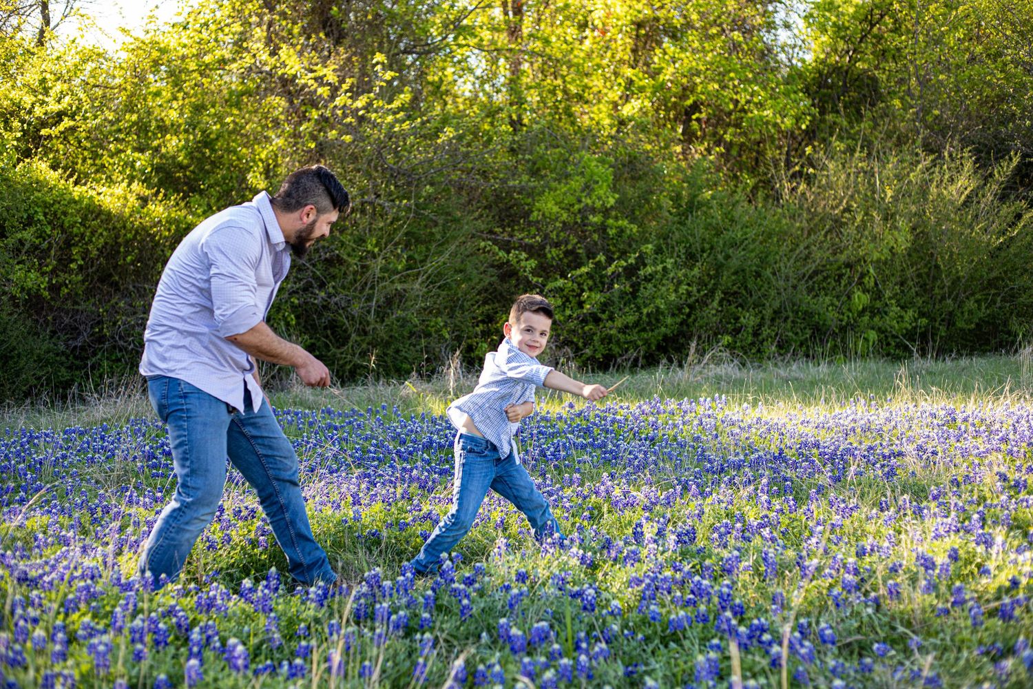 A father and young son play together in a vibrant Texas bluebonnet field surrounded by lush green trees.