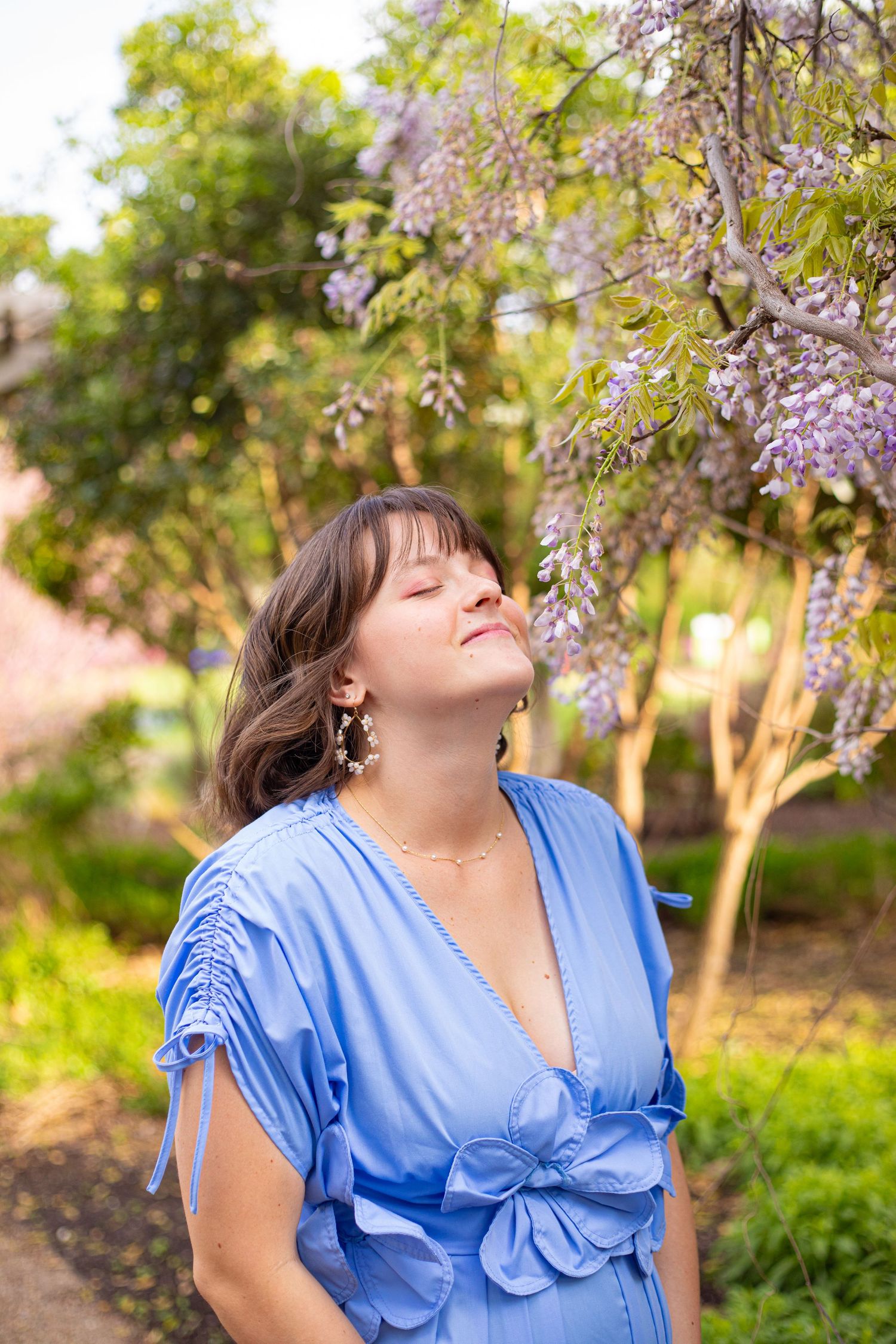 Woman in blue dress poses under blooming wisteria flowers during spring photo shoot.