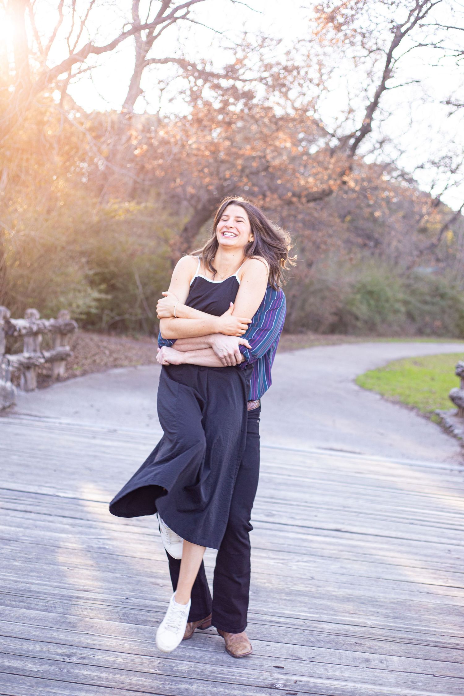 A person in a black dress twirls playfully on a wooden boardwalk at sunset.