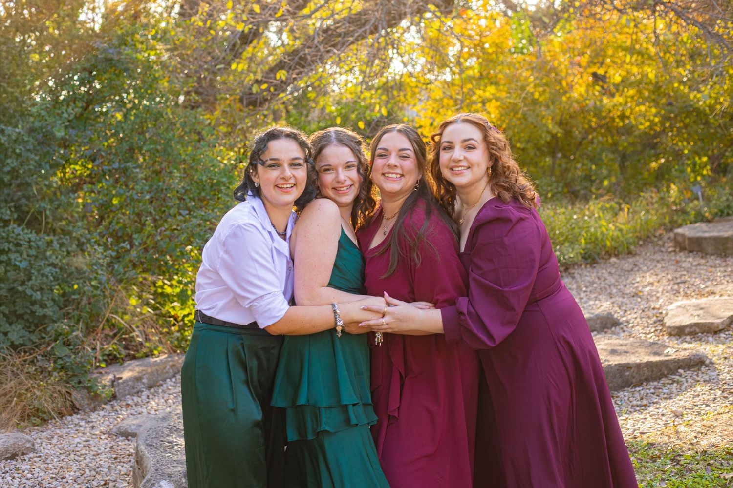 Four young women in elegant dresses smile together outdoors among autumn foliage in a warm golden light.