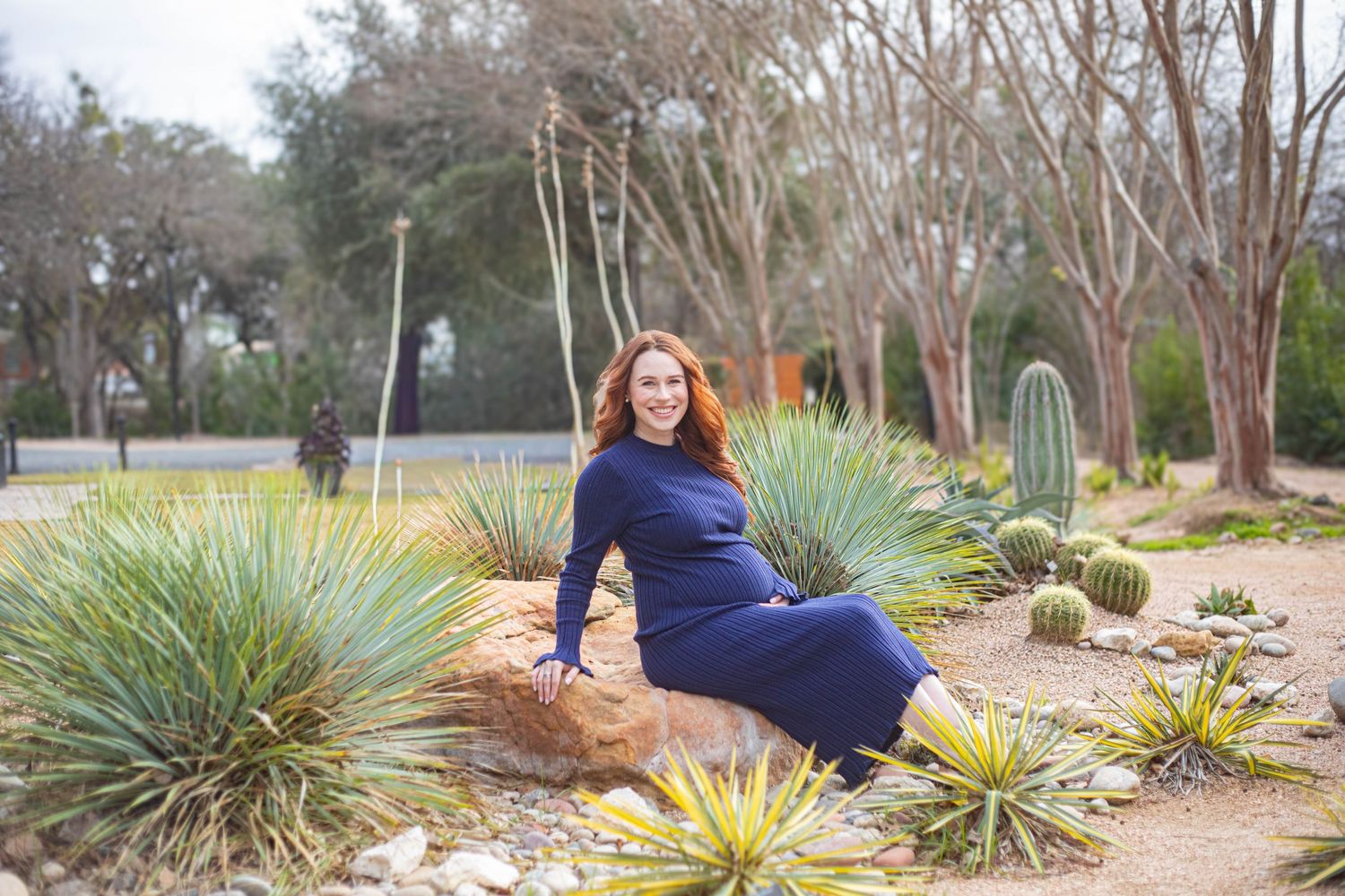 Woman in blue dress sitting on rock among cacti and yuccas in a desert garden with trees in background.
