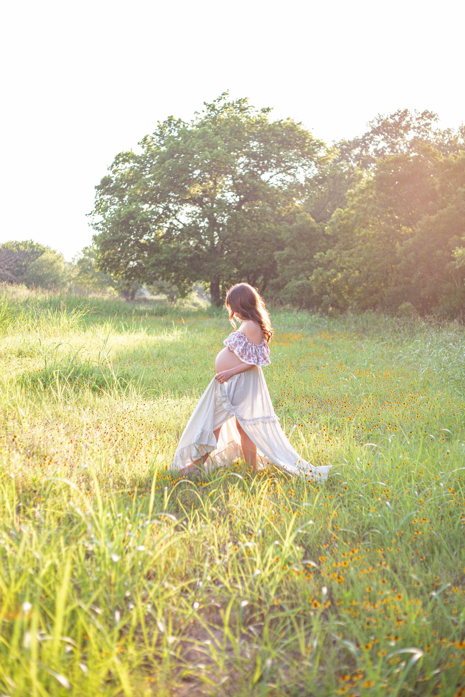 Pregnant woman in white dress walking through sunny green field, cradling baby bump, with trees in background.
