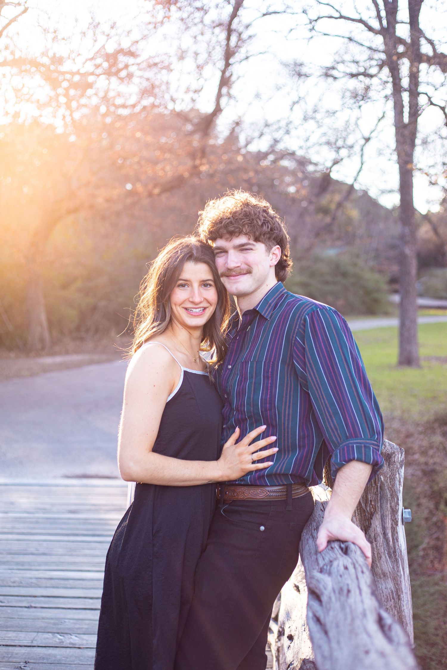 A couple poses together on a wooden walkway with warm sunset lighting in the background.