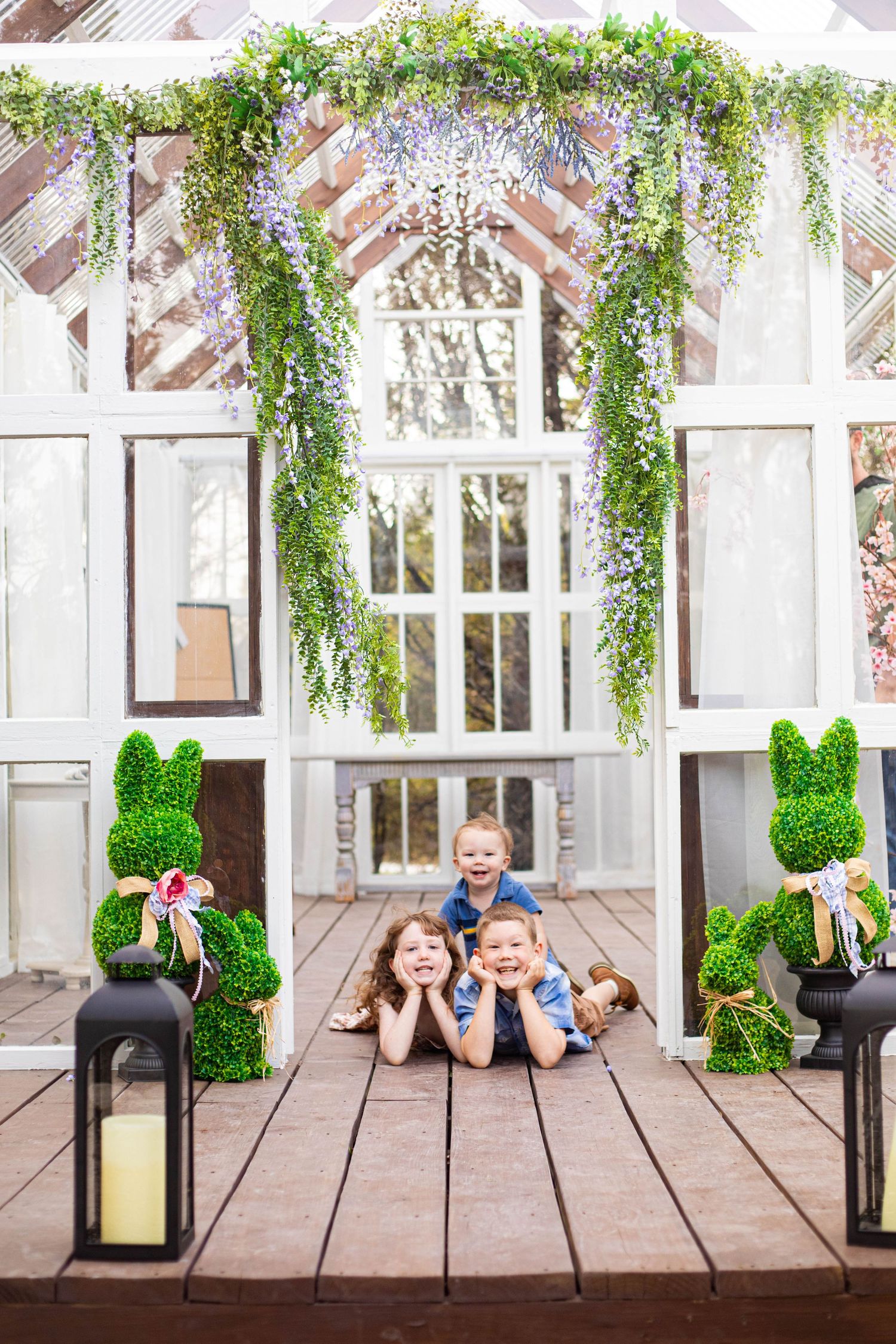 Three happy children lying on a wooden deck surrounded by Easter bunny topiaries and floral arch decorations.