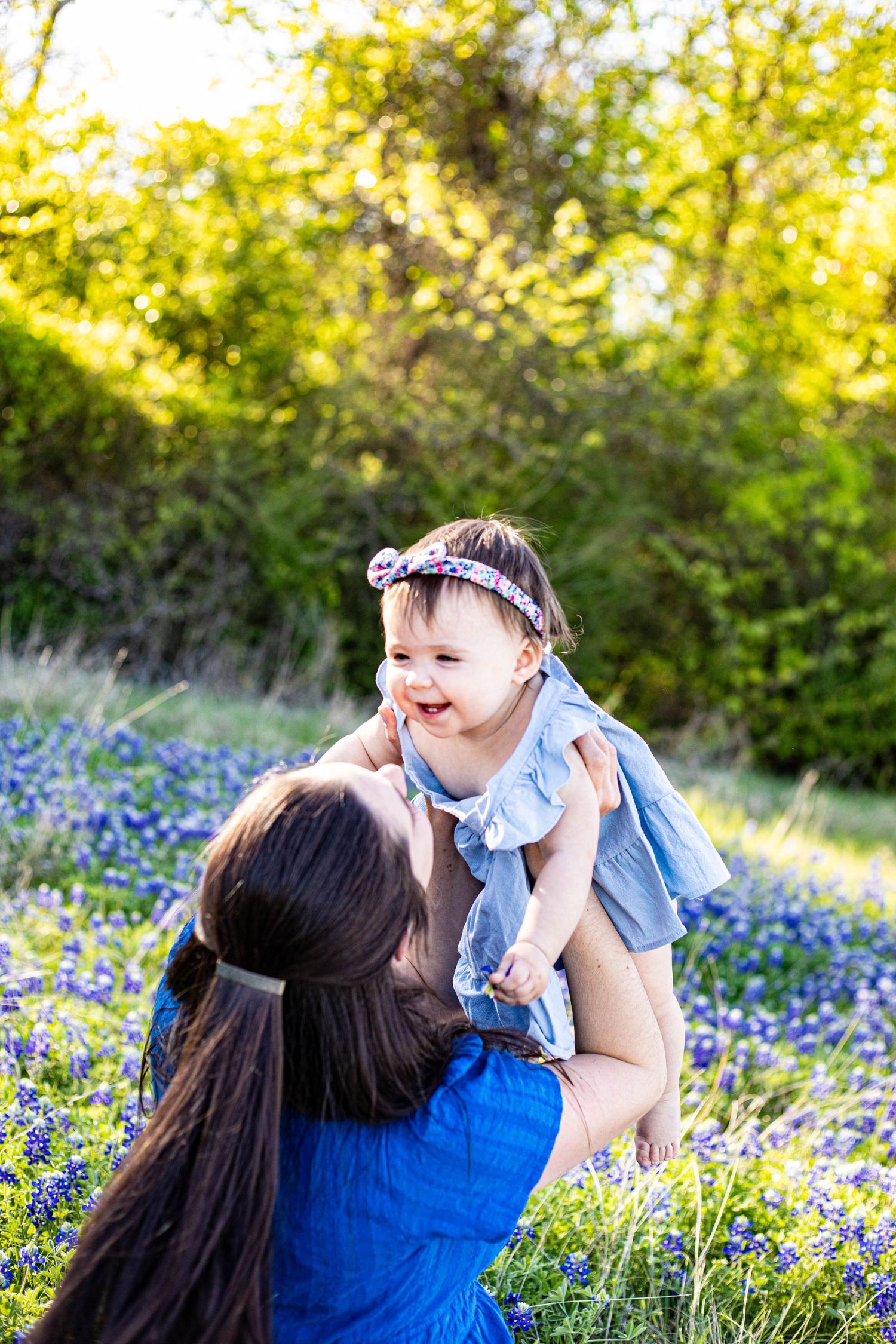 A mother lifts her laughing baby girl in a field of bluebonnets, both wearing blue dresses outdoors.