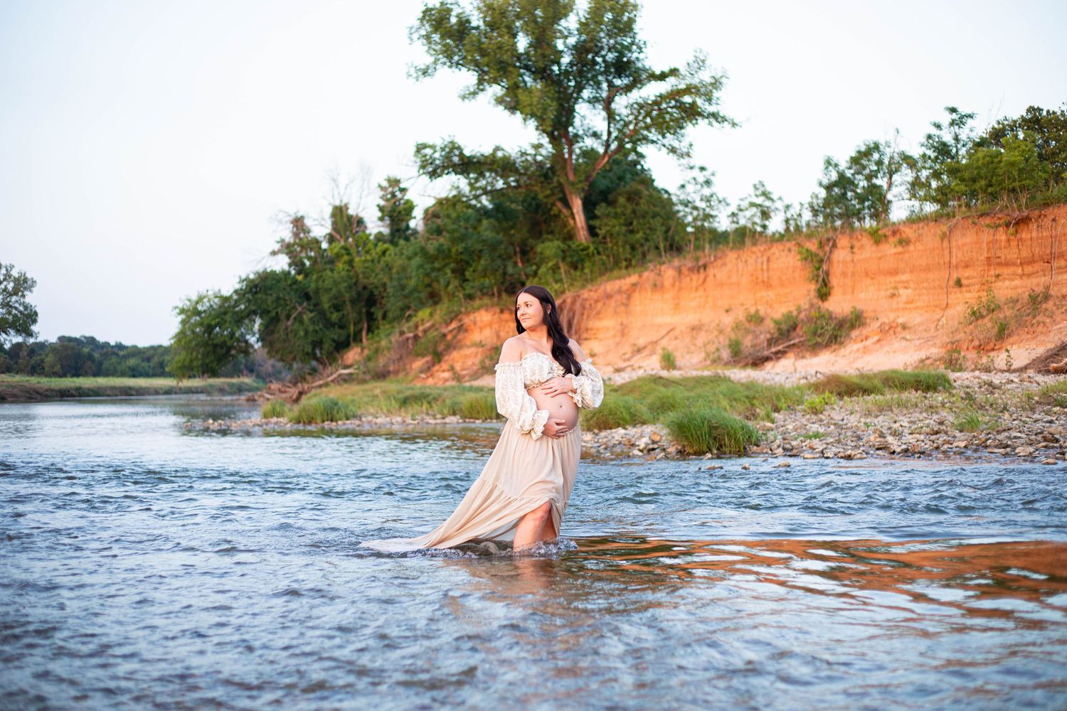 Pregnant woman in flowing beige dress wading in a river with red clay banks and green trees in background.