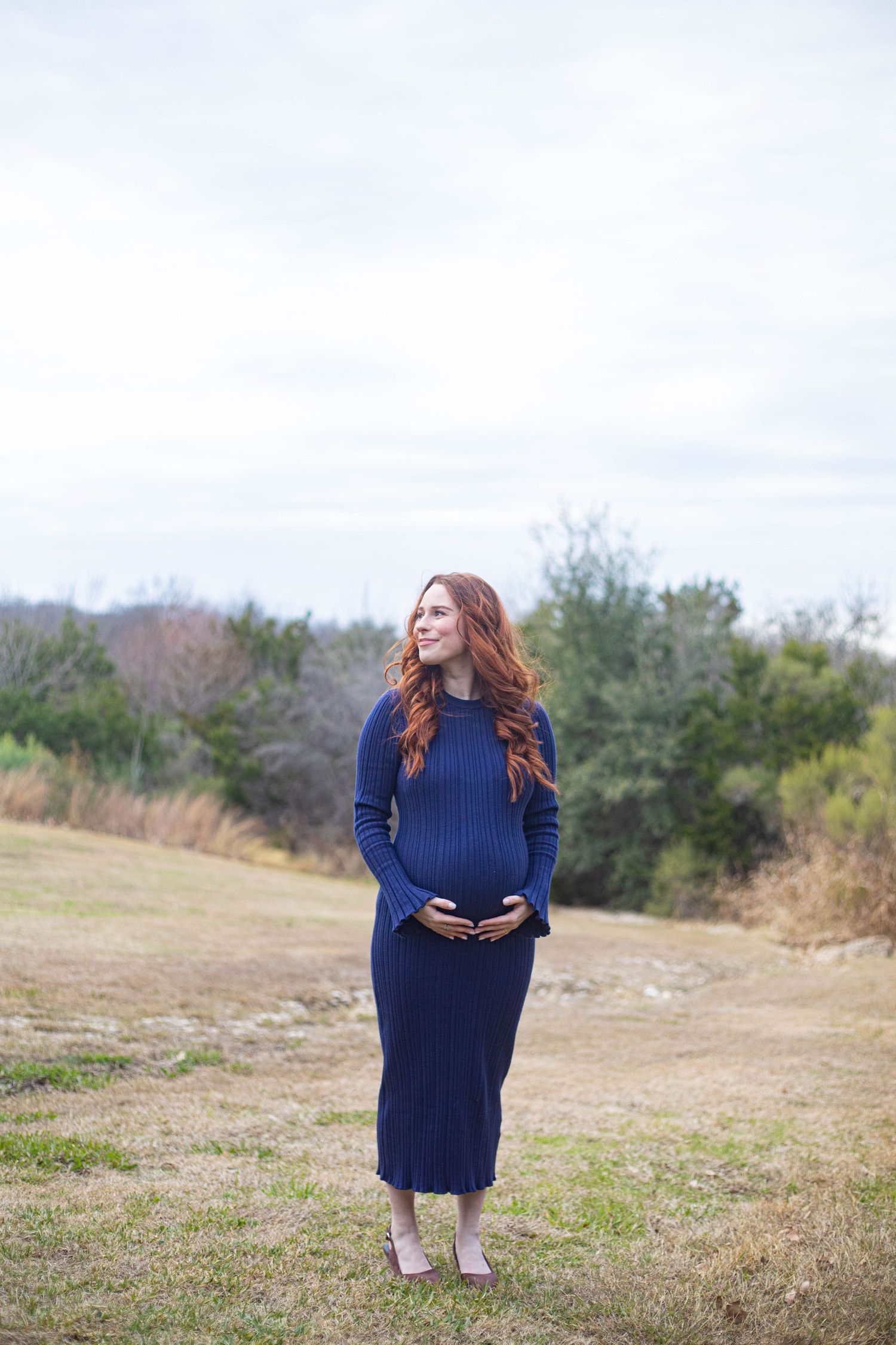 Pregnant woman in navy ribbed dress cradling baby bump outdoors in a grassy field with trees behind her.