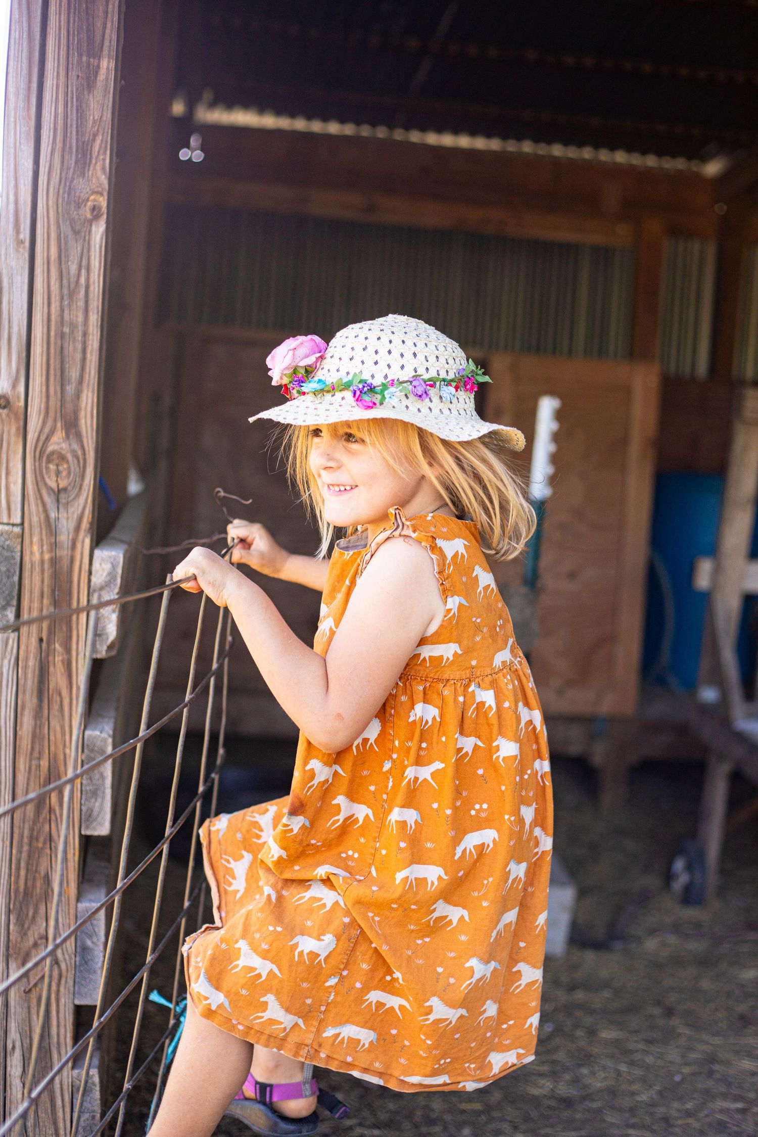 Child wearing an orange dress with horse print and sun hat leaning against farm fence.