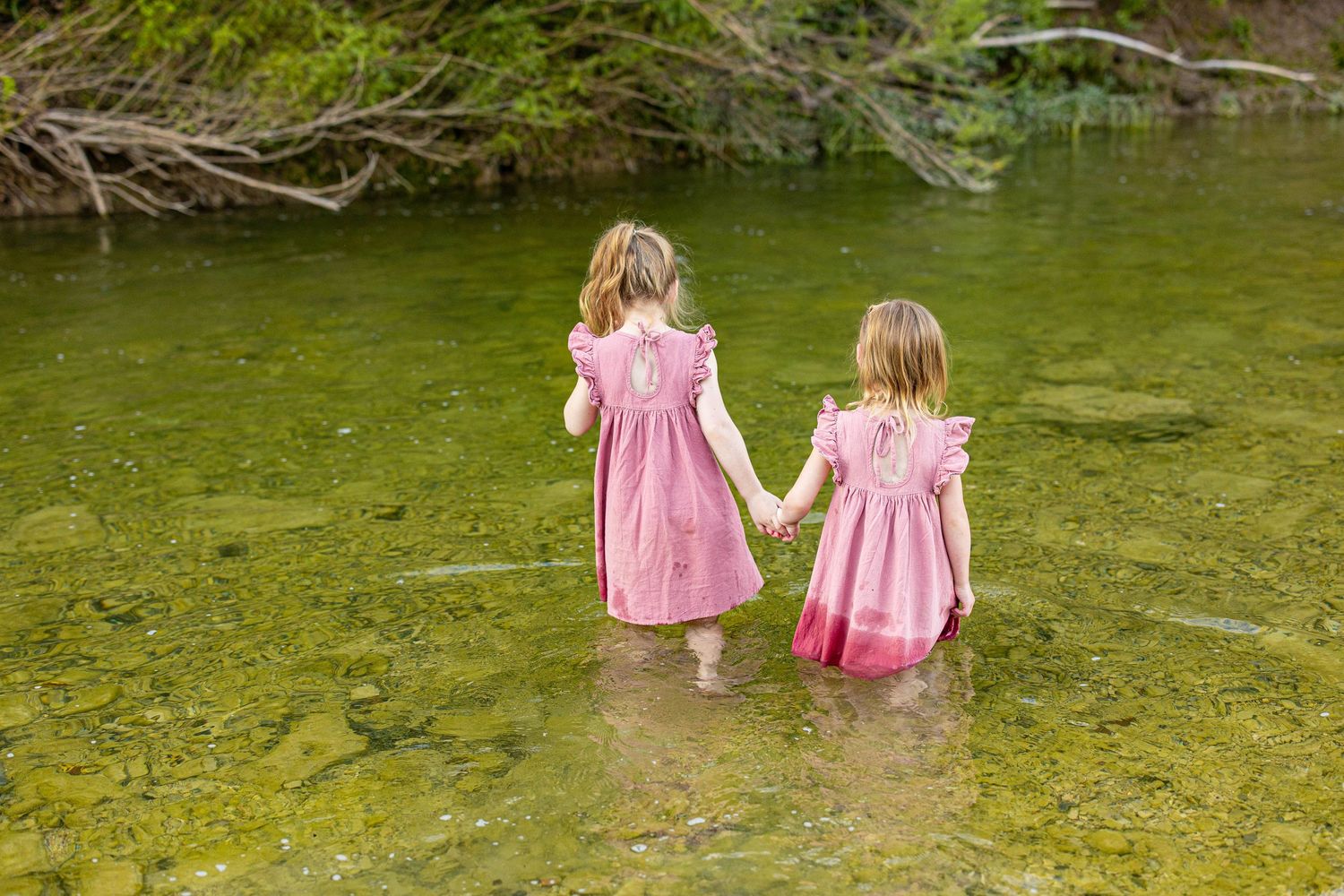Two figures in matching pink dresses wade hand-in-hand through shallow green water.
