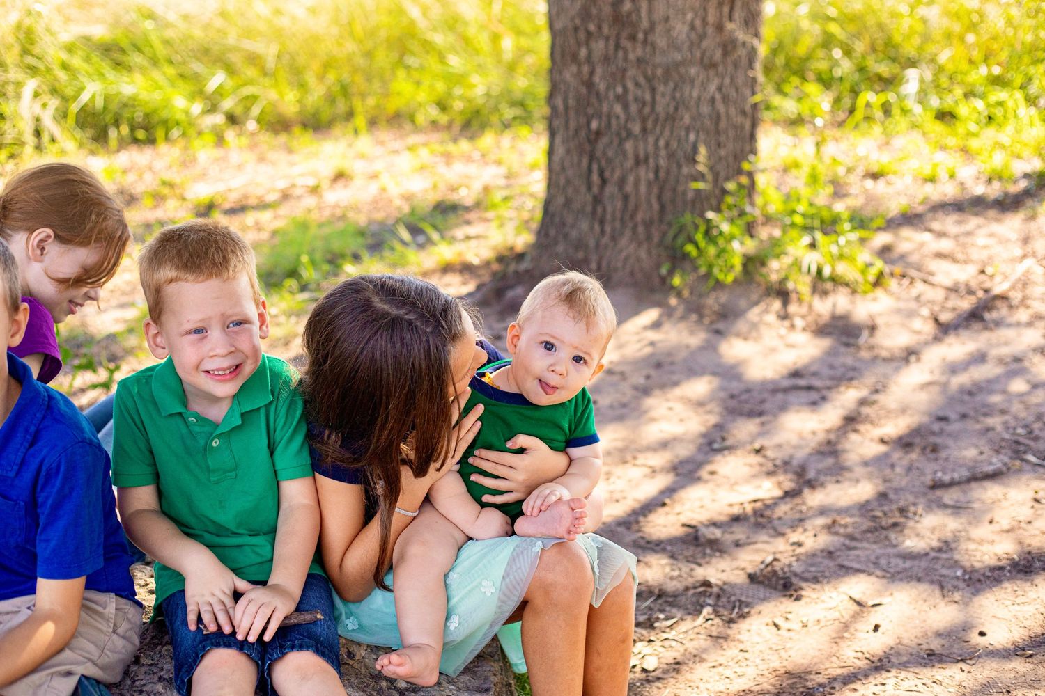 Family group sitting together under a large tree in a sunny outdoor setting.