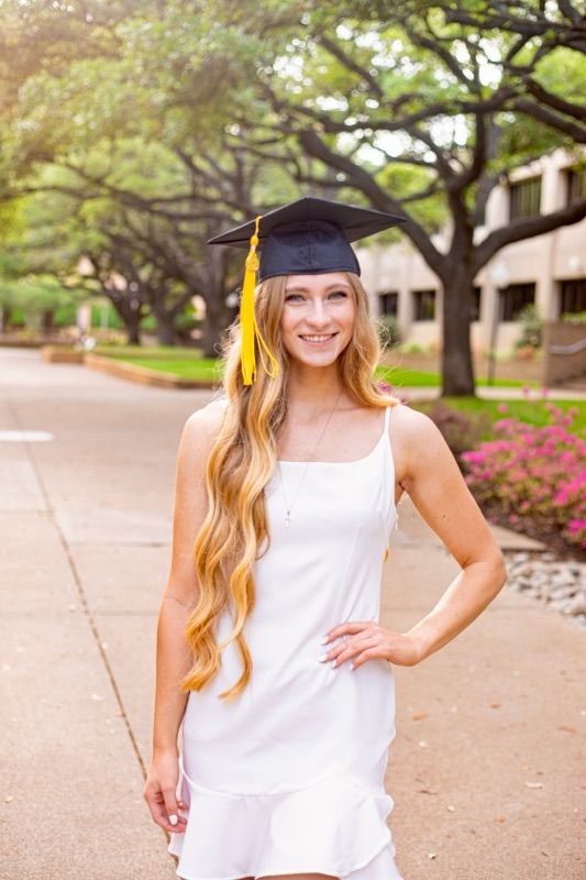Graduate in white dress and cap poses on tree-lined campus walkway.