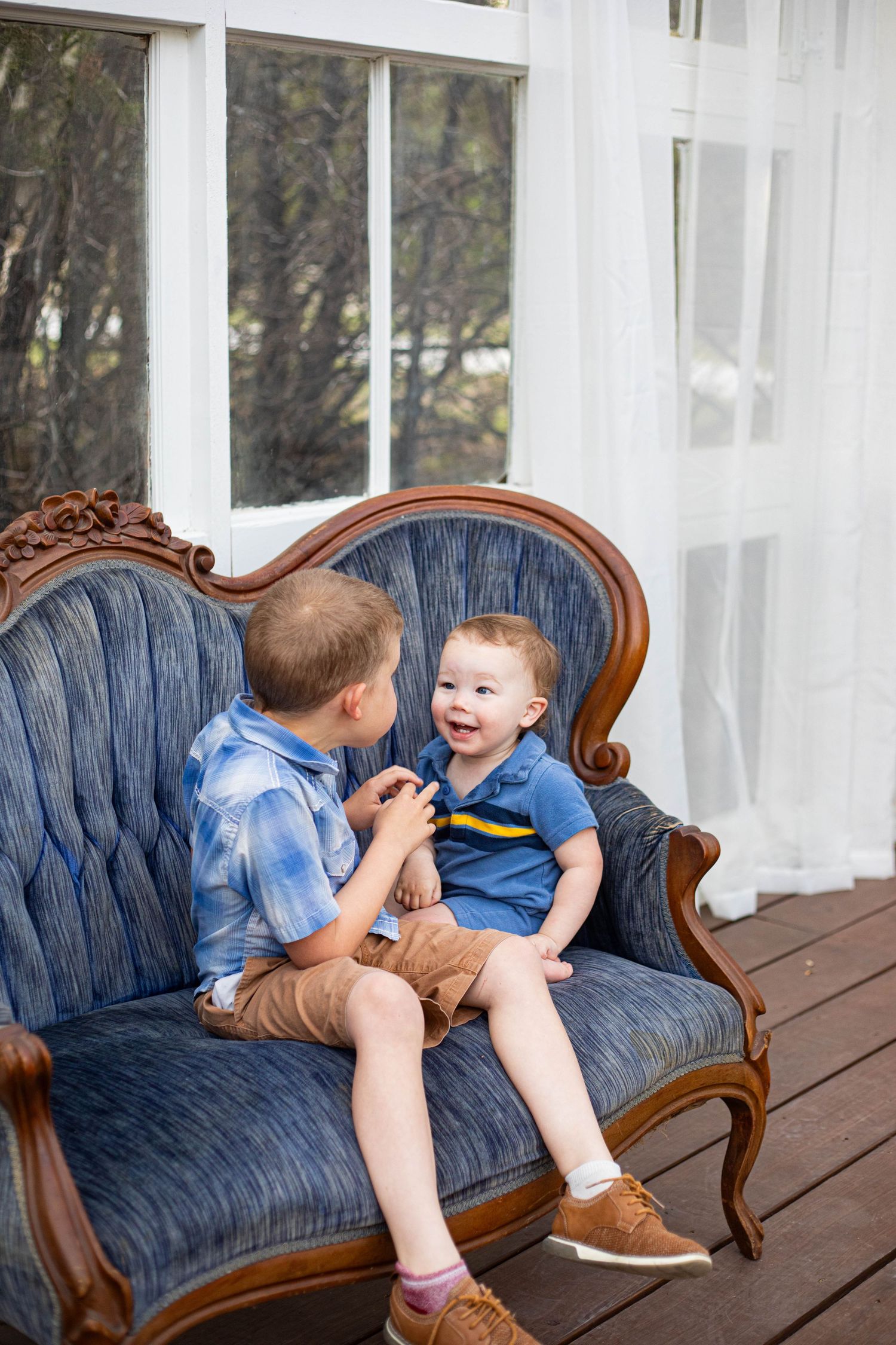 Two young boys sitting together on a vintage blue velvet sofa, sharing a joyful moment indoors.
