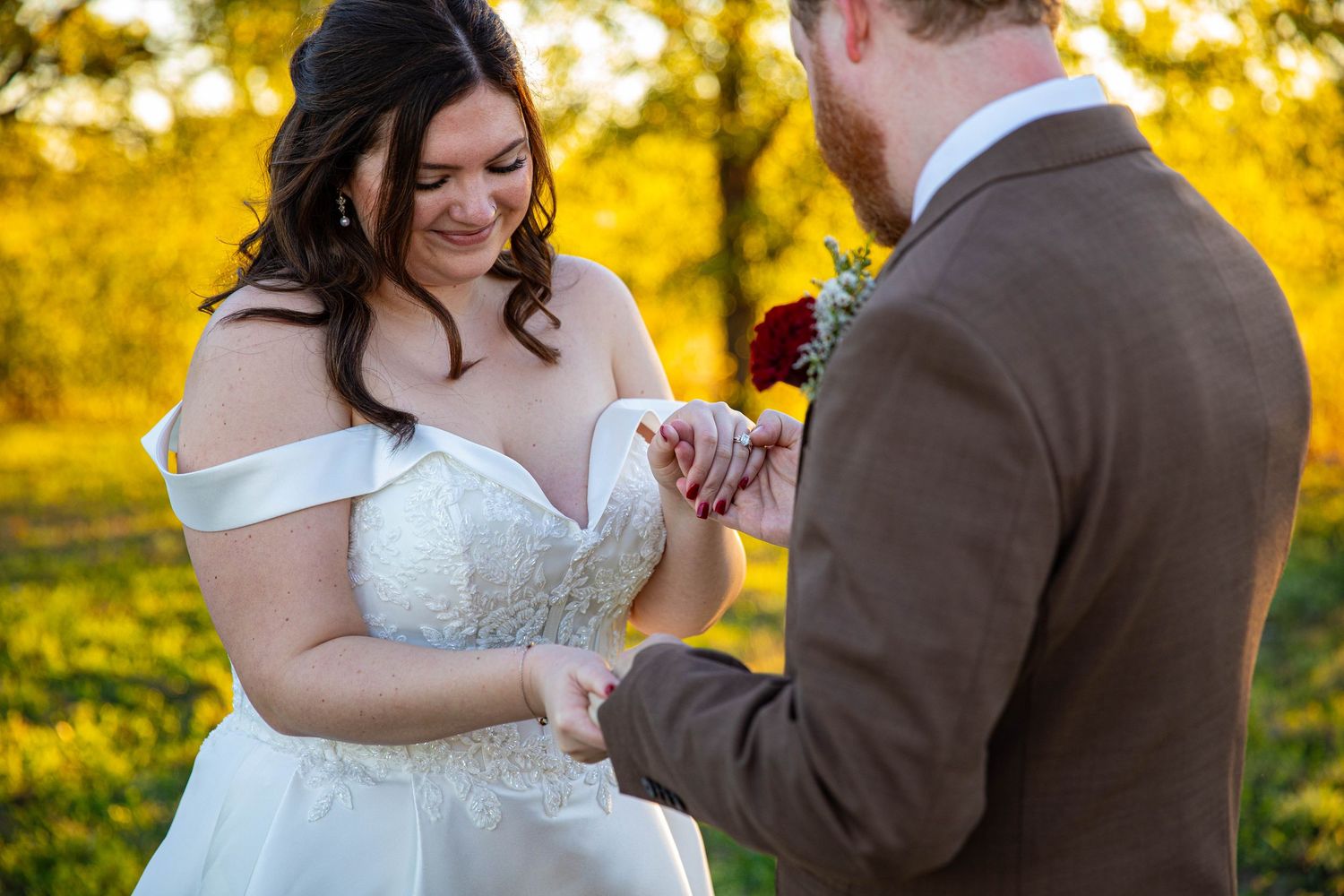 Bride placing wedding ring on groom's finger outdoors during golden hour ceremony, wearing off-shoulder lace gown.