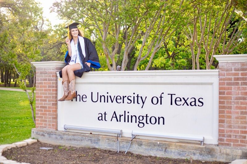 Graduate sits on University of Texas at Arlington entrance sign in cap and gown.