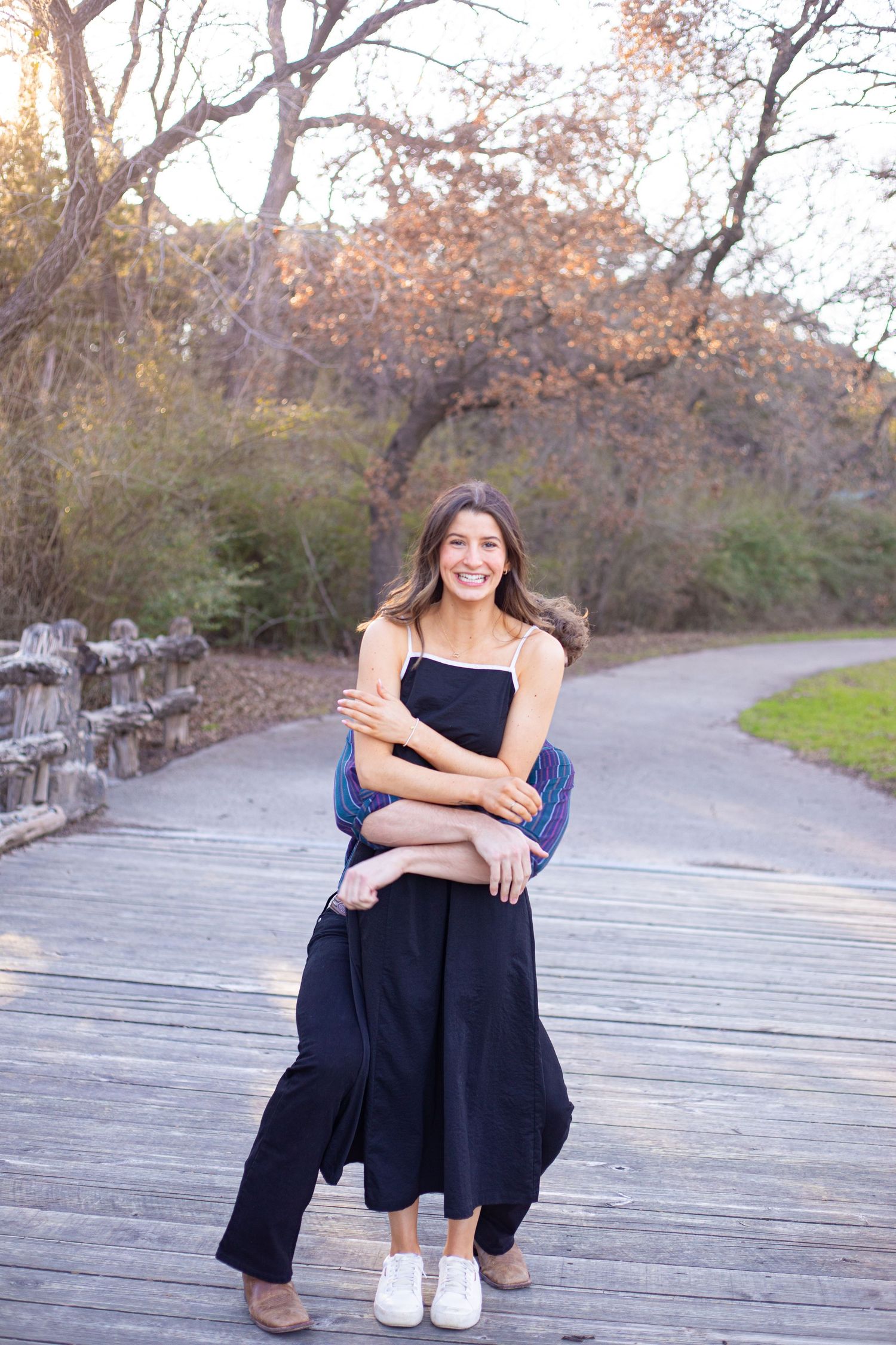 A person in a black dress sits casually on a wooden boardwalk surrounded by autumn trees.
