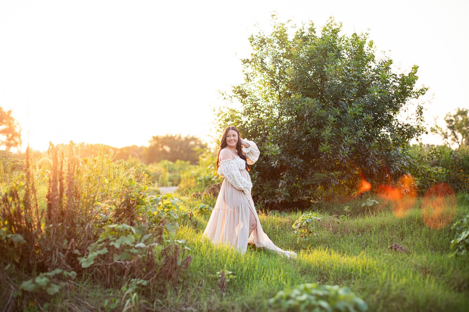 Pregnant woman in flowing dress standing in sunlit garden at golden hour, surrounded by lush green plants and trees.