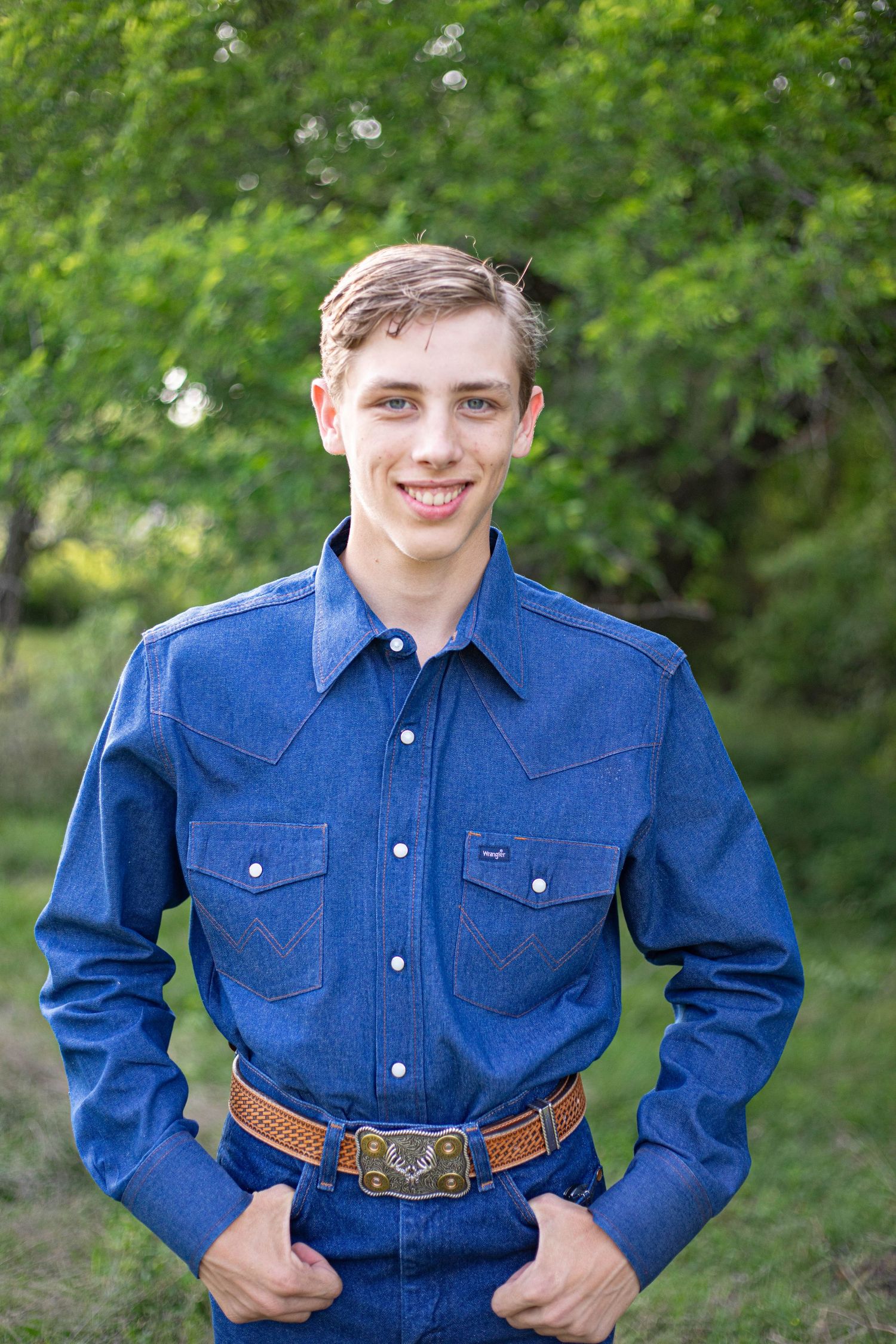 Smiling teenage boy in denim Wrangler shirt and decorative belt buckle posing outdoors with green trees behind him.