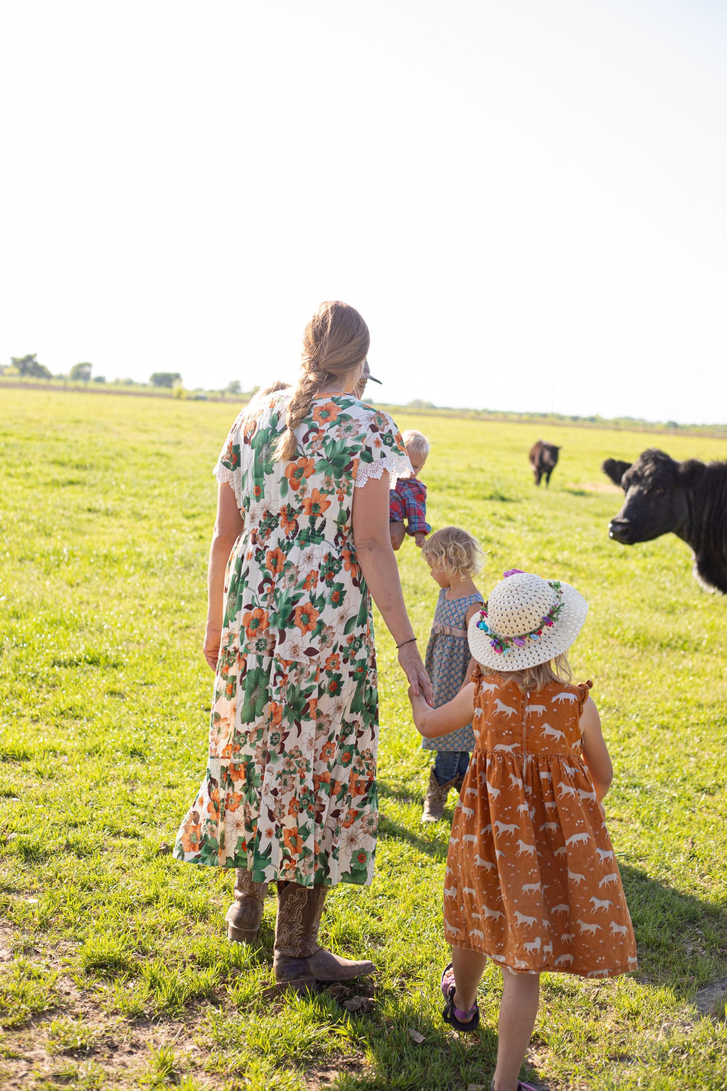 Two people in floral dresses walking through a grassy pasture with cattle in the background.