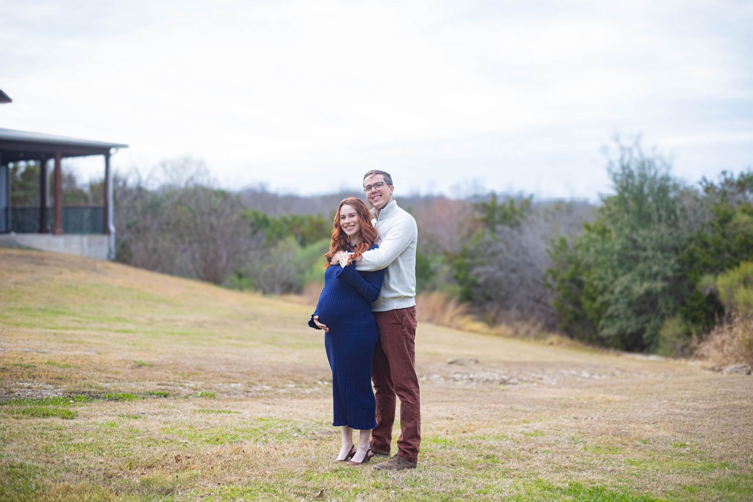 Couple embracing outdoors in a field with trees and cloudy sky, woman in navy dress, man in gray sweater.
