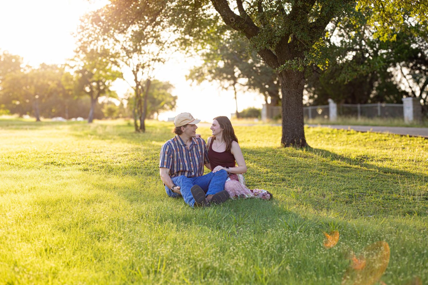 Couple sitting together on lush green grass under a tree during golden hour sunset in a park.