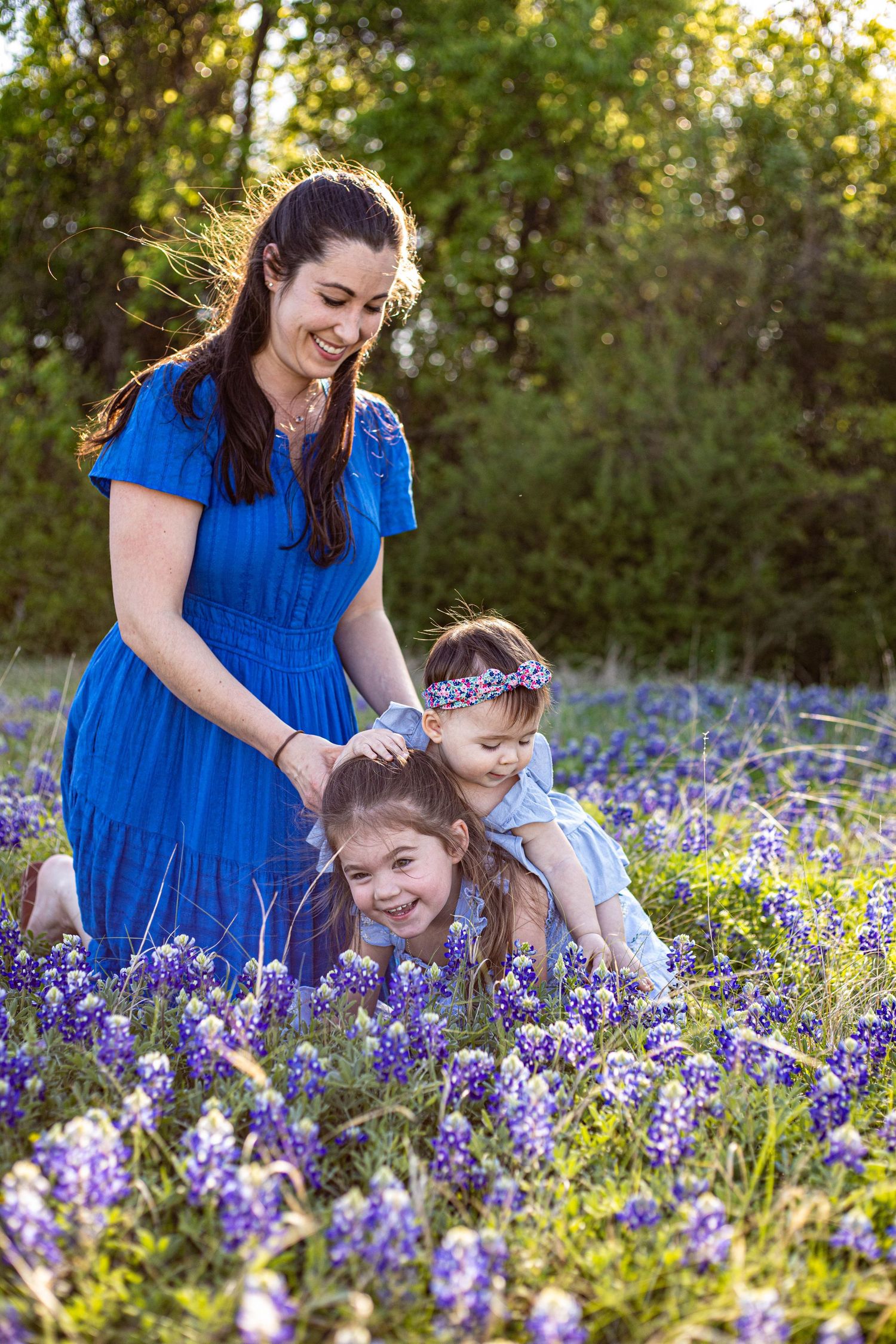 A smiling mother in a blue dress plays with her two young daughters in a field of purple bluebonnets.
