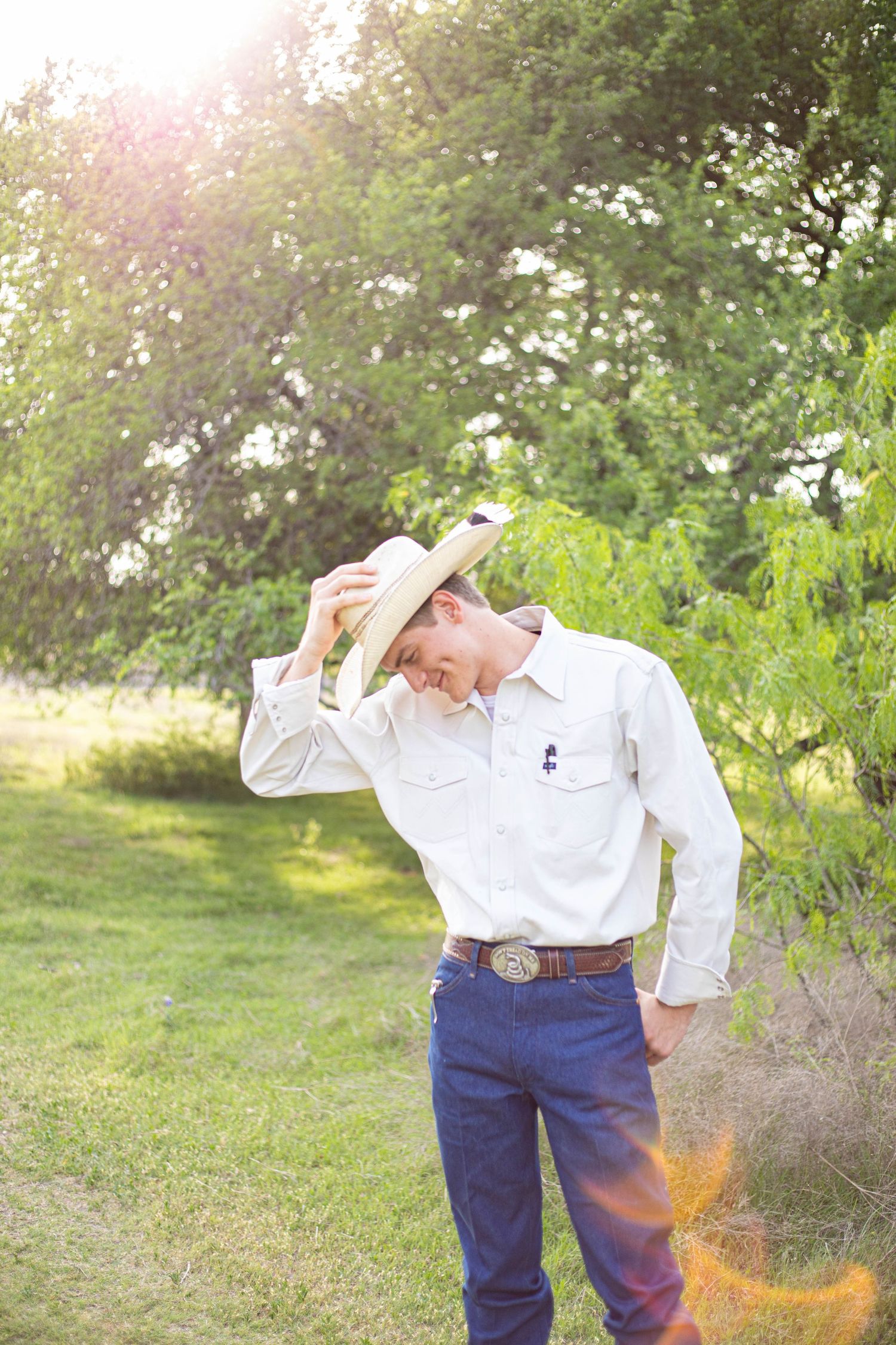 Young man in white western shirt tipping his cowboy hat outdoors in a sunny green landscape.