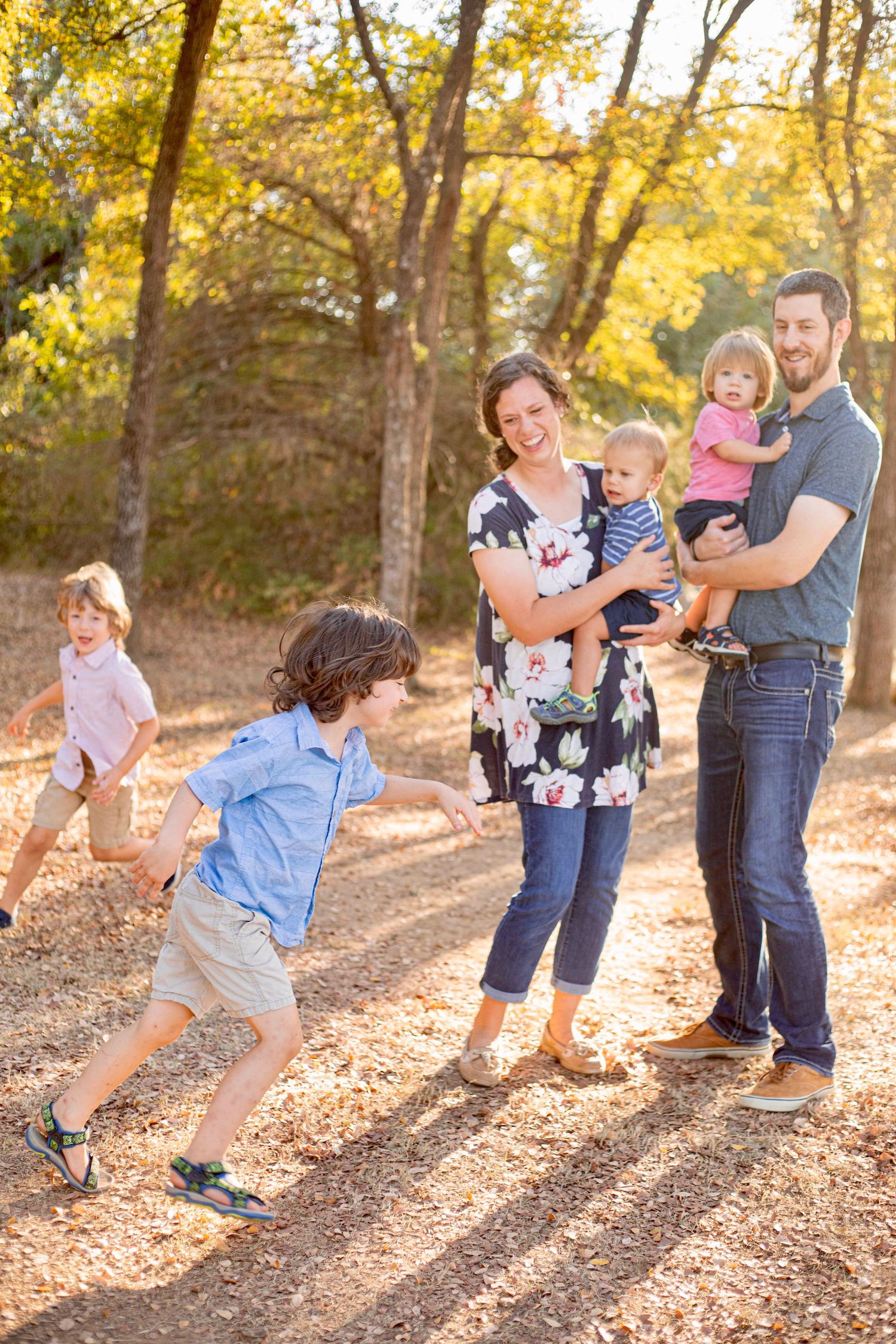 A family enjoys a playful autumn photoshoot in a sun-dappled forest setting with golden leaves.
