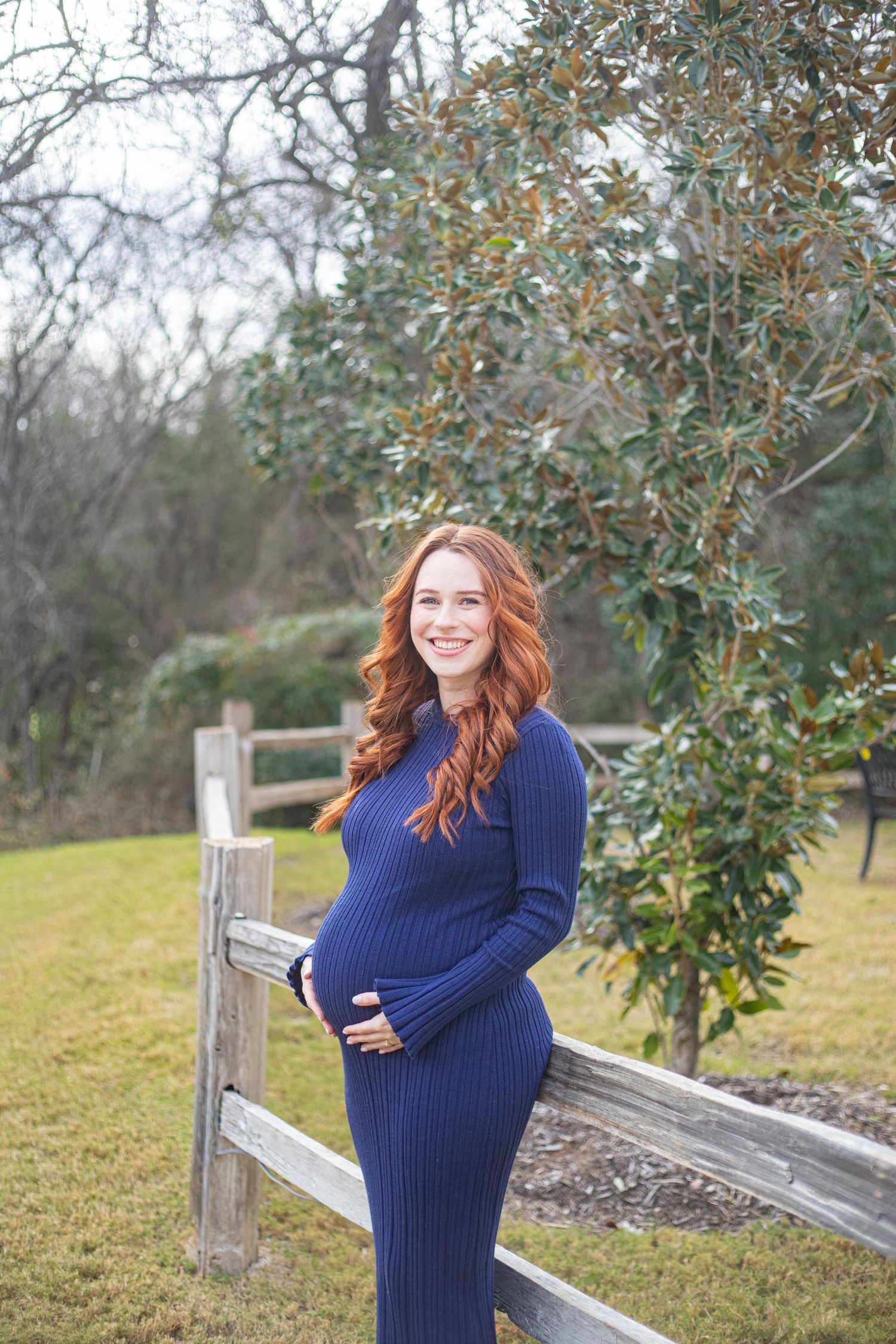 Smiling pregnant woman in navy blue ribbed dress holding baby bump outdoors by wooden fence.