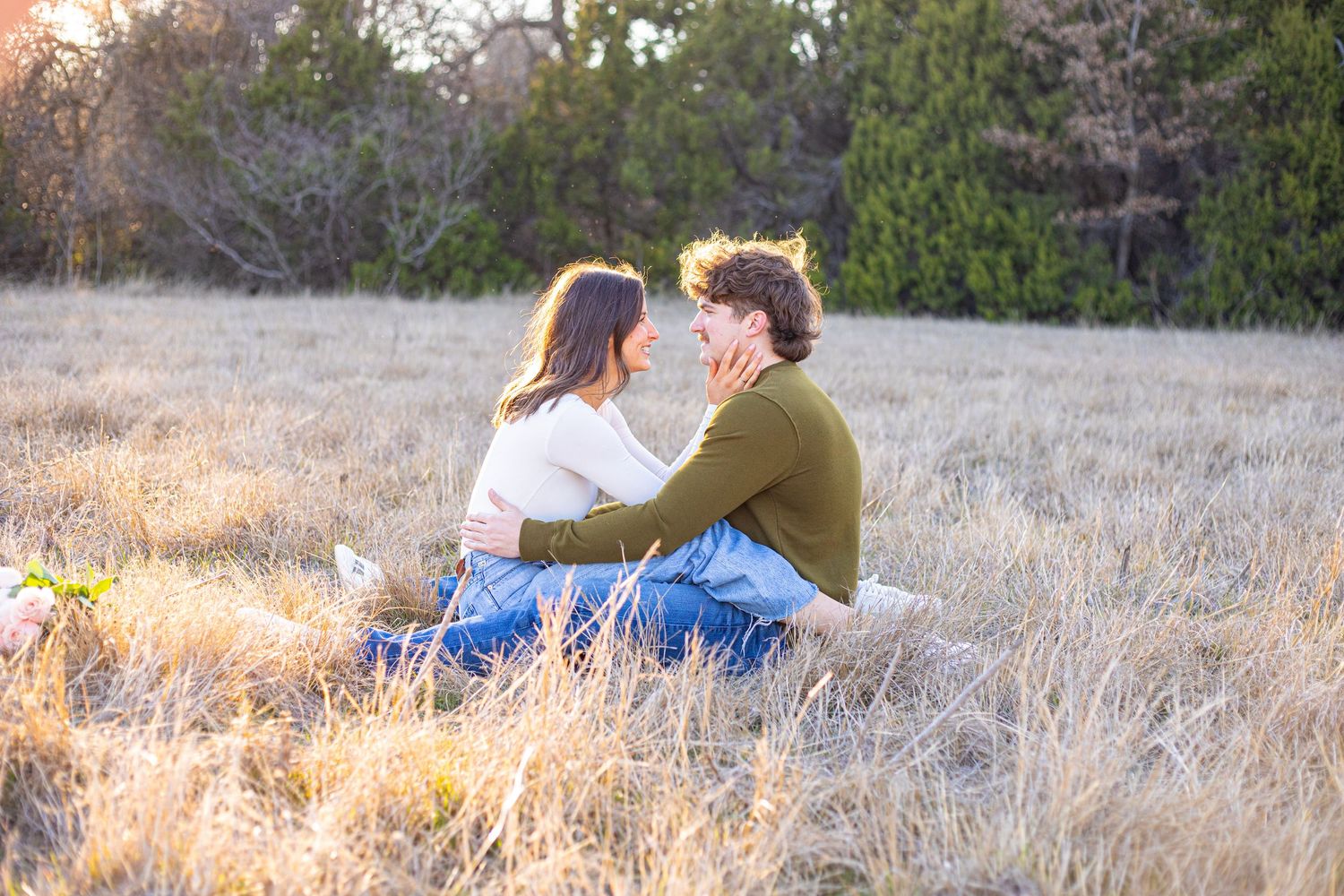 Couple laying in tall grass during sunset with trees visible in the distance.