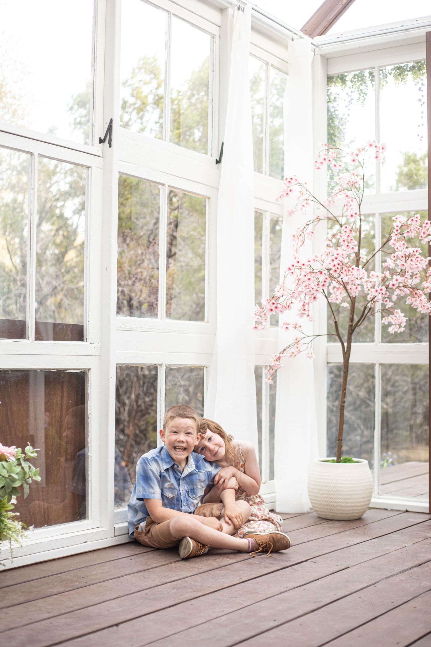 Two children sitting together on a wooden deck in a bright sunroom with large windows and a pink cherry blossom tree.