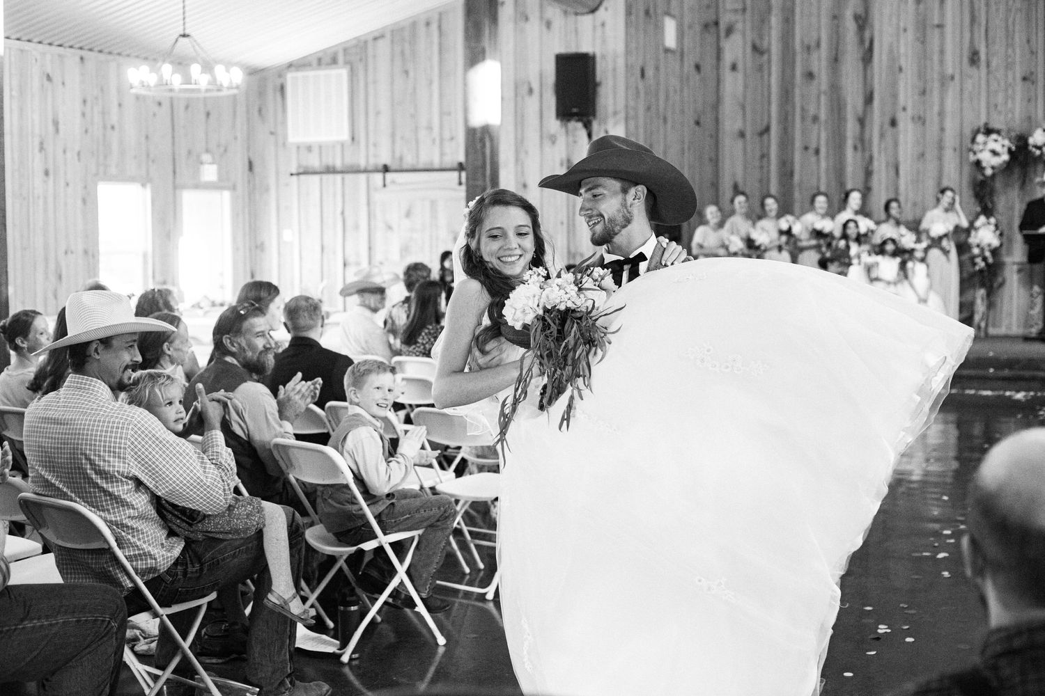 Wedding guests seated in rows of white chairs inside a rustic wooden barn venue during ceremony.