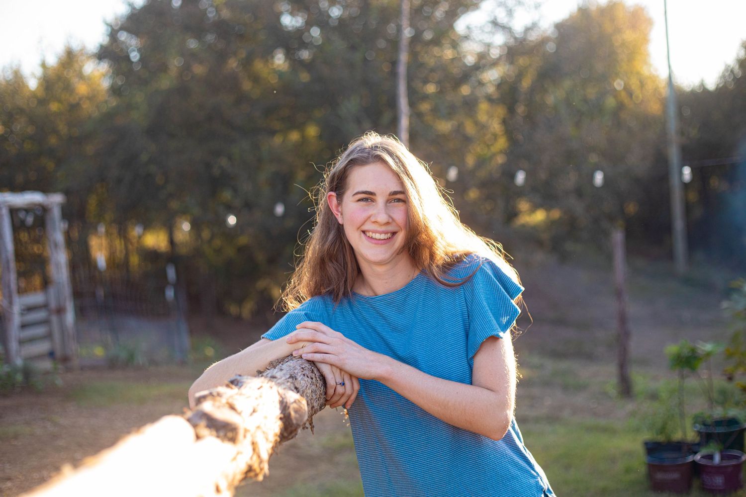 Person in casual blue top leaning against wooden log in backyard setting.