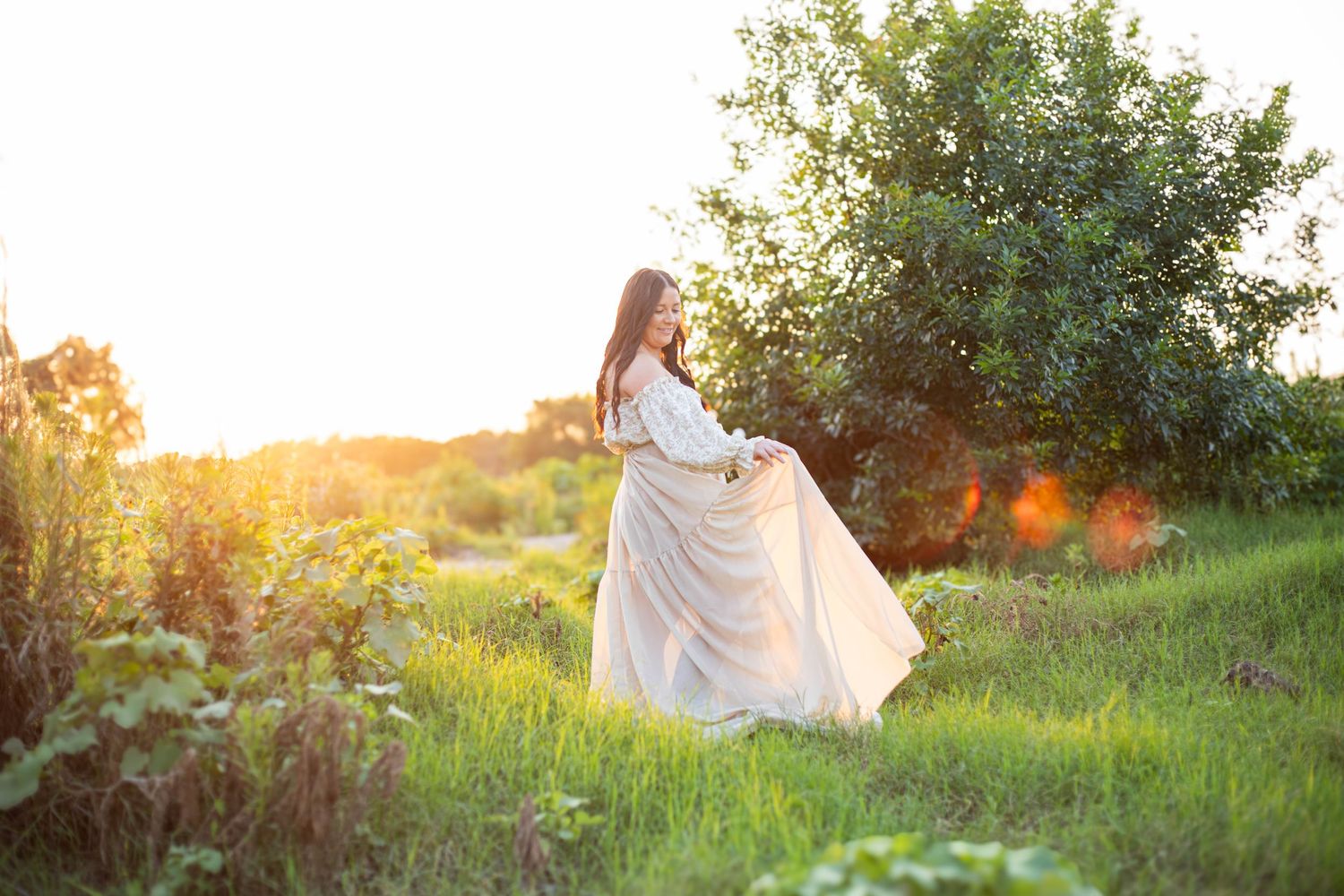 A woman in a flowing white dress twirls in a sunlit green meadow with trees in the background.