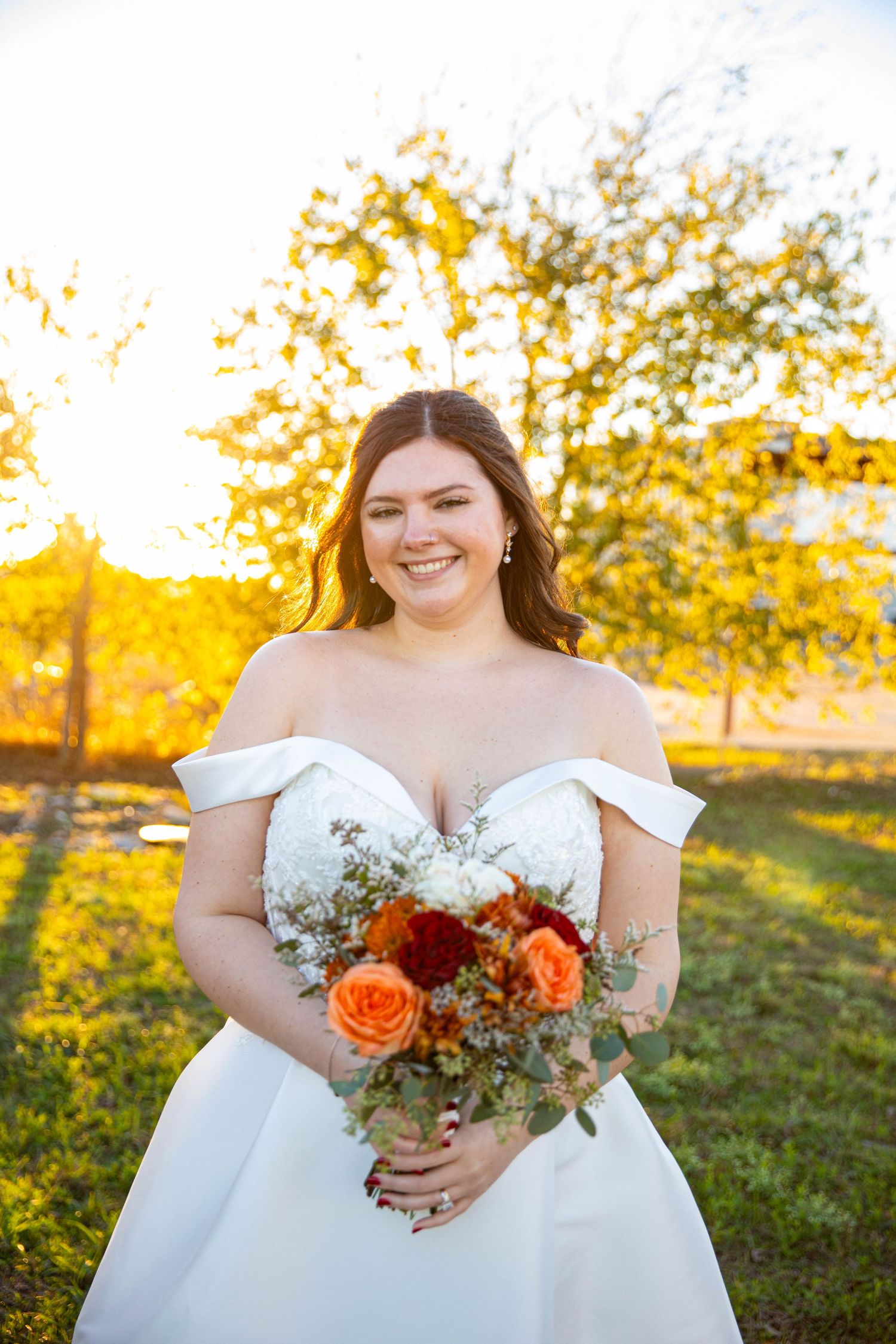 Smiling bride in off-shoulder white gown holds orange and white bouquet outdoors during golden hour sunset.