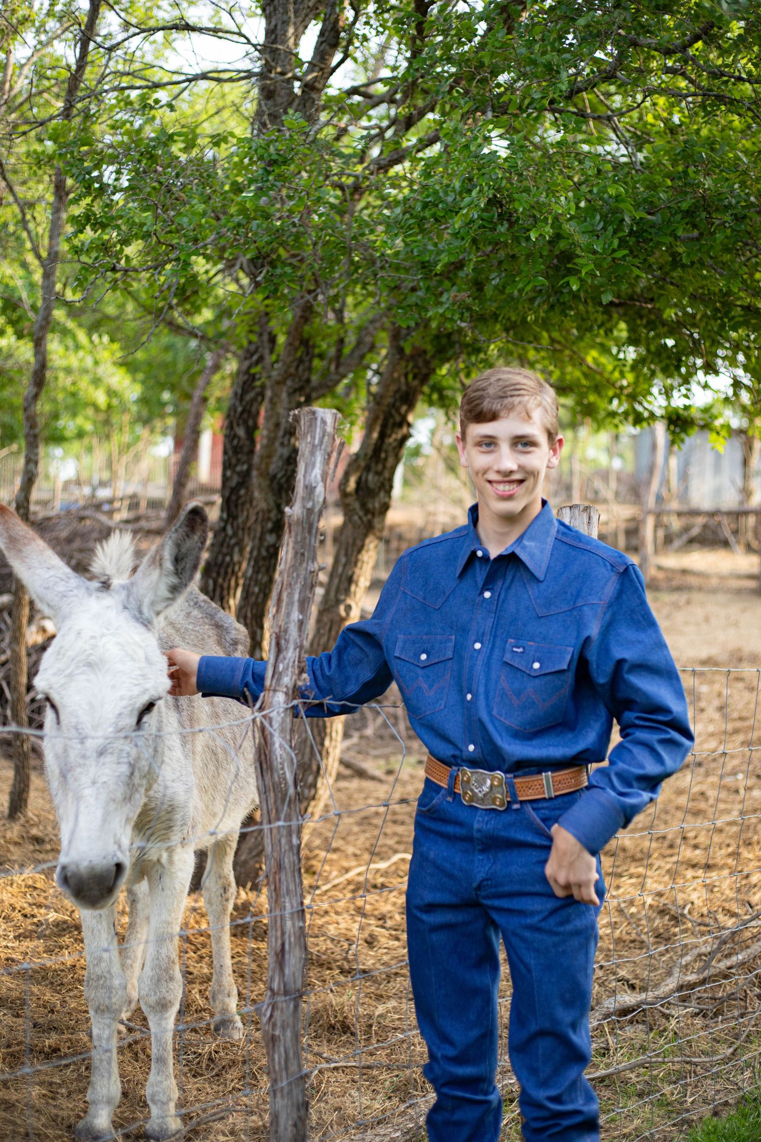 Young man in blue denim outfit smiling beside a white donkey near a fence with green trees in background.