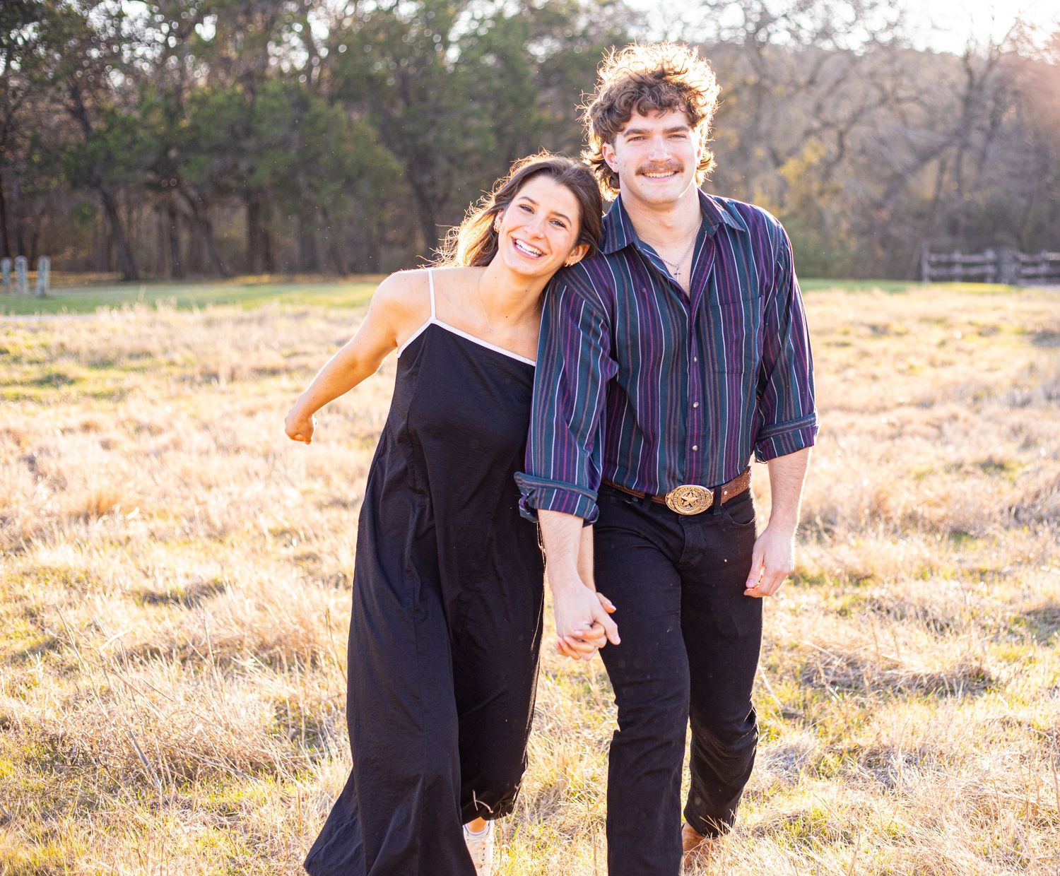 A couple walks hand in hand through a sunlit field with joyful expressions.