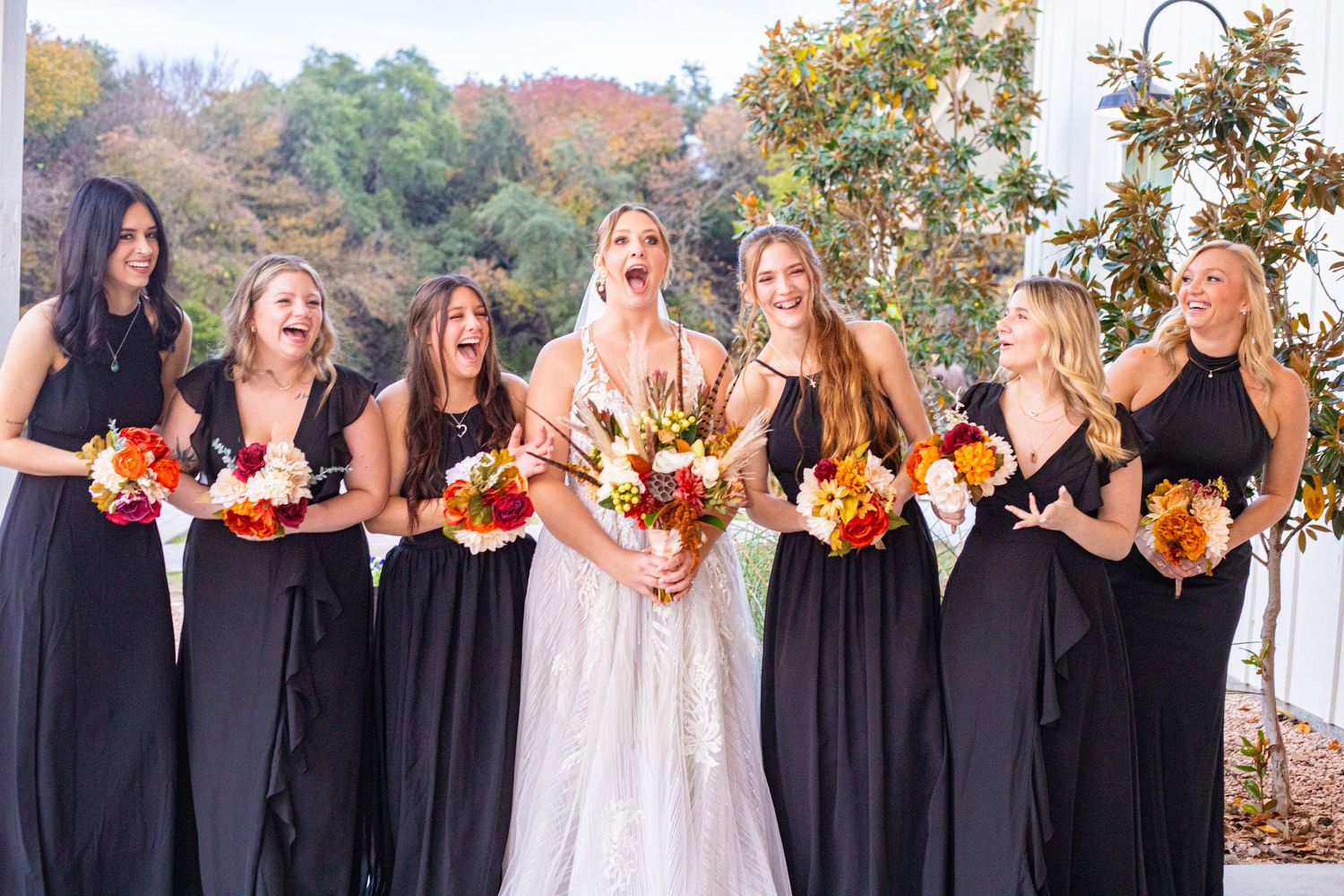 Bridal party with bridesmaids in black dresses holding autumn-colored bouquets outdoors.