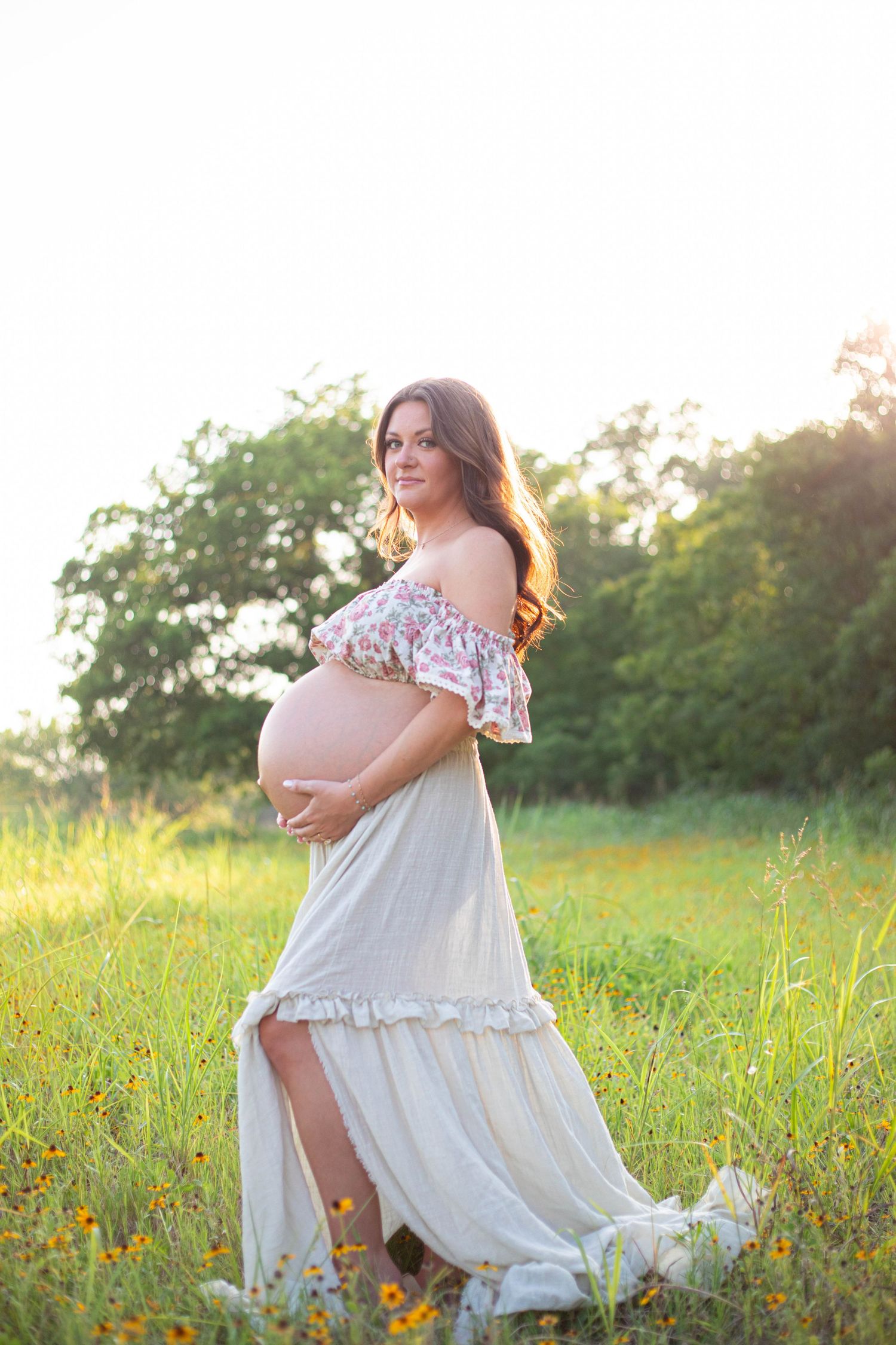 Pregnant woman in floral off-shoulder top and white maxi skirt cradling baby bump in sunlit meadow.
