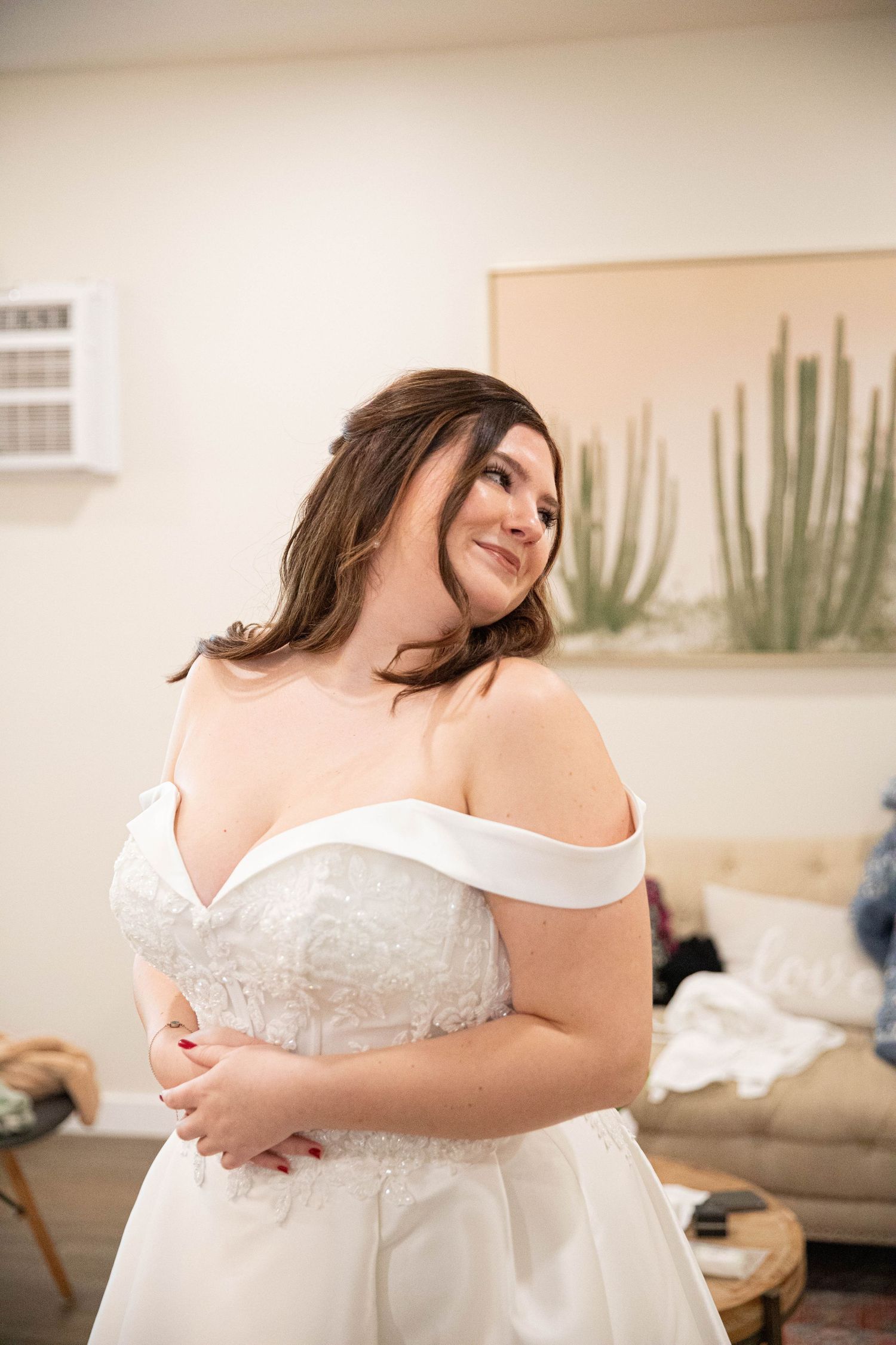Bride in an off-shoulder white lace wedding gown smiling softly in a bridal suite with cactus wall art.