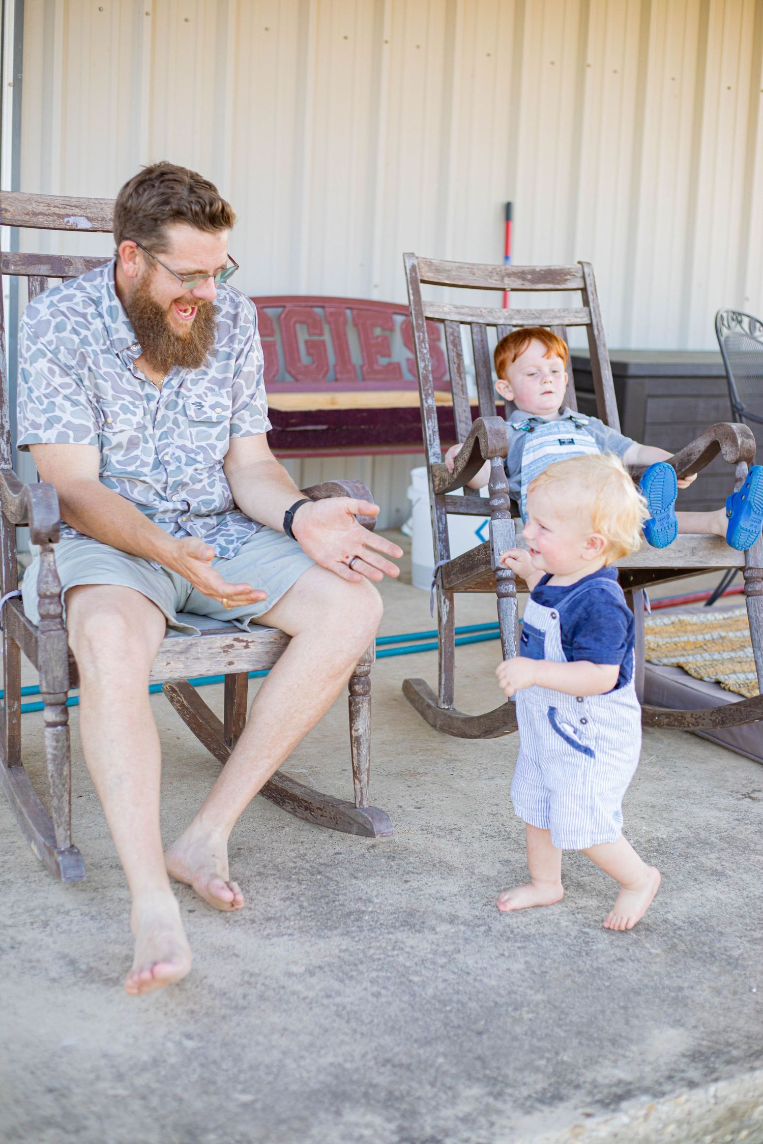 Bearded father sitting in rocking chair on porch smiling at two young redhead toddler boys playing outdoors.