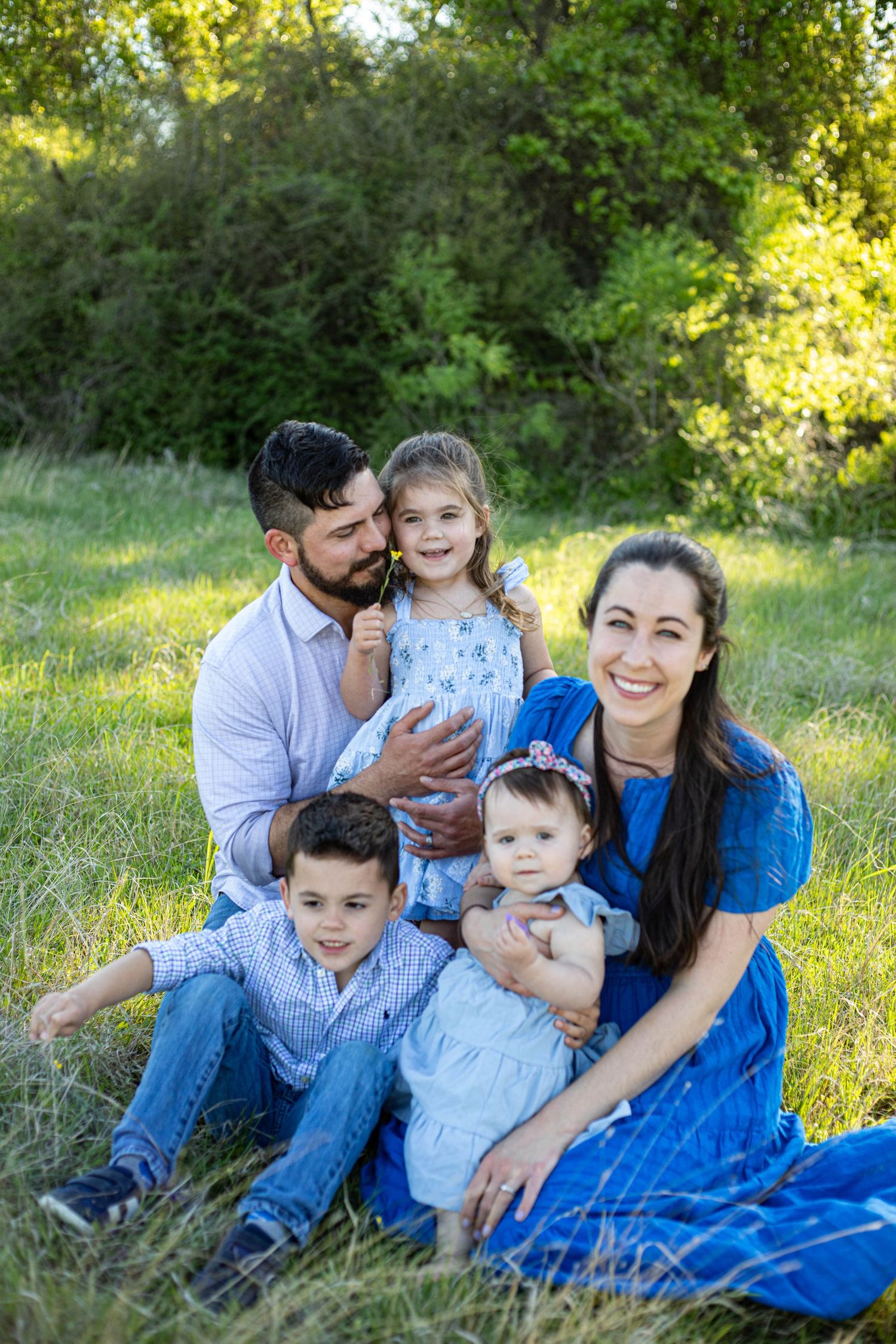 Happy family of five sitting in green grass outdoors, parents with two young children and a baby in blue outfits.